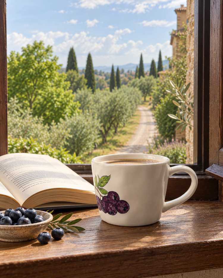 Hand-painted mug with wild blueberry and leaf details displayed by a sunlit Tuscan-style window, blending nature-inspired design with everyday ceramics.