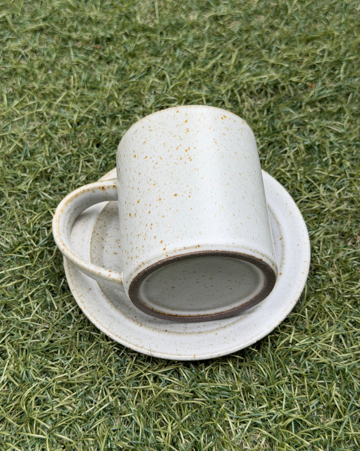 A speckled off-white ceramic mug and matching saucer placed on green grass.