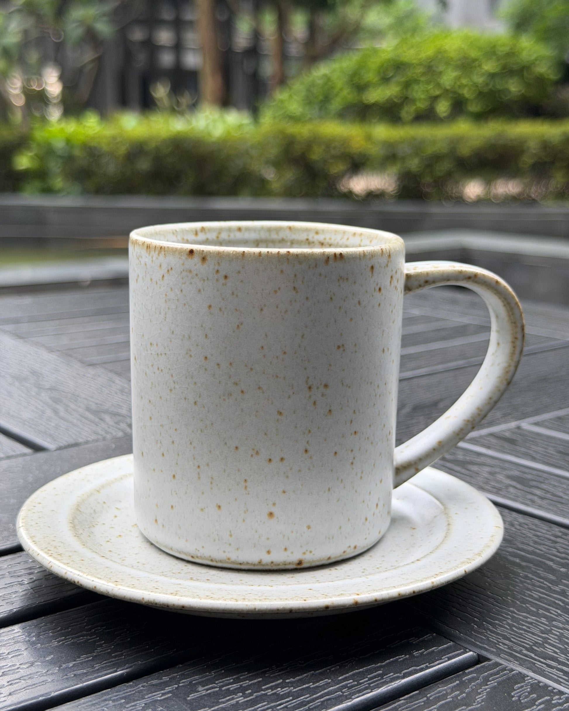 A speckled off-white ceramic mug with a matching saucer, placed on a dark gray wooden table with a blurred outdoor garden background.