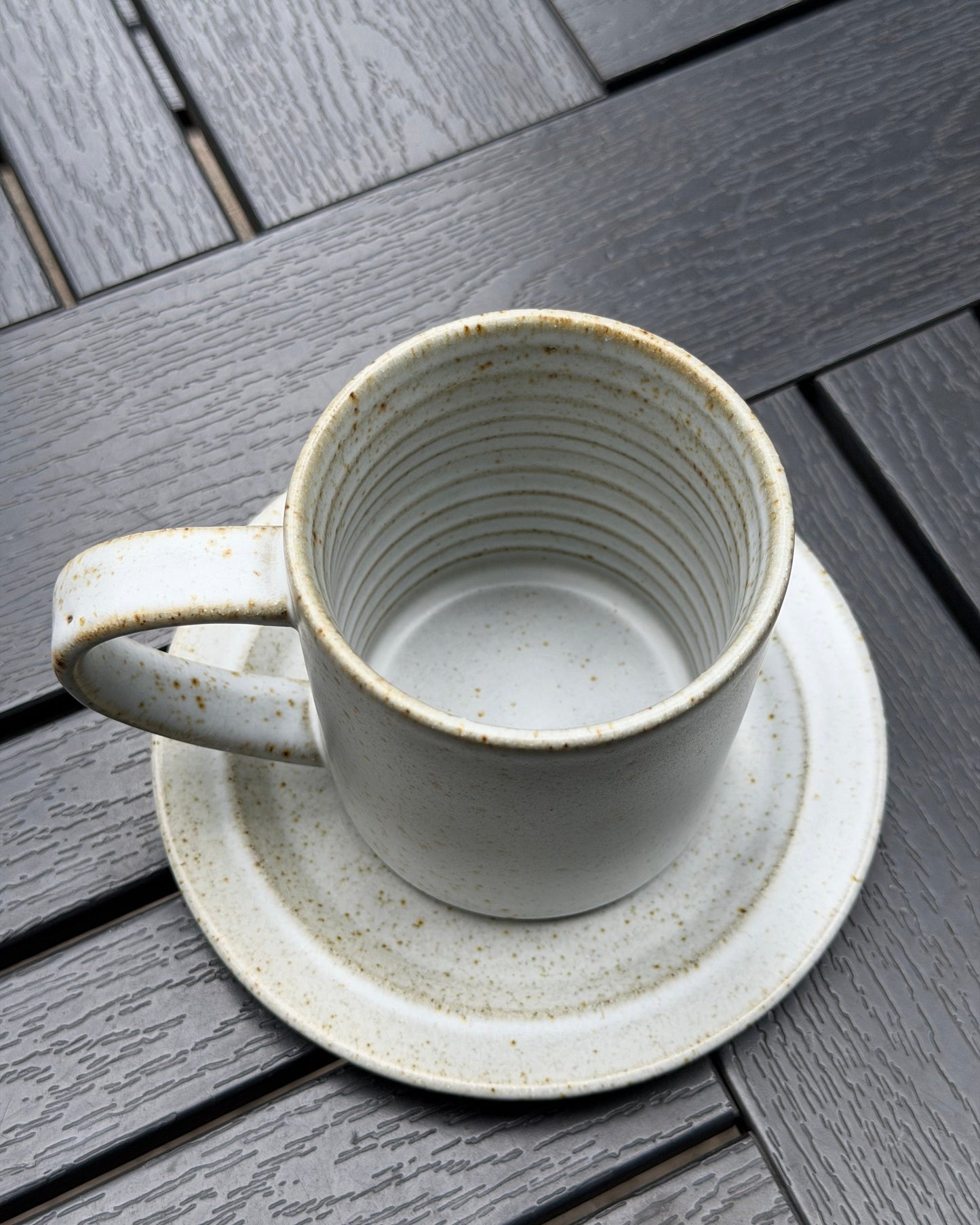 A speckled stoneware mug with matching saucer, photographed from above on a dark gray wooden slatted surface.