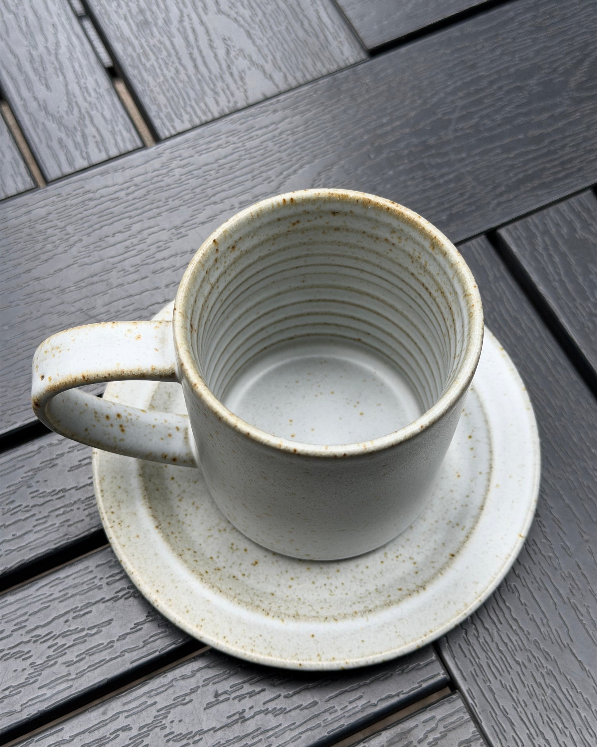 A speckled stoneware mug with matching saucer, photographed from above on a dark gray wooden slatted surface.