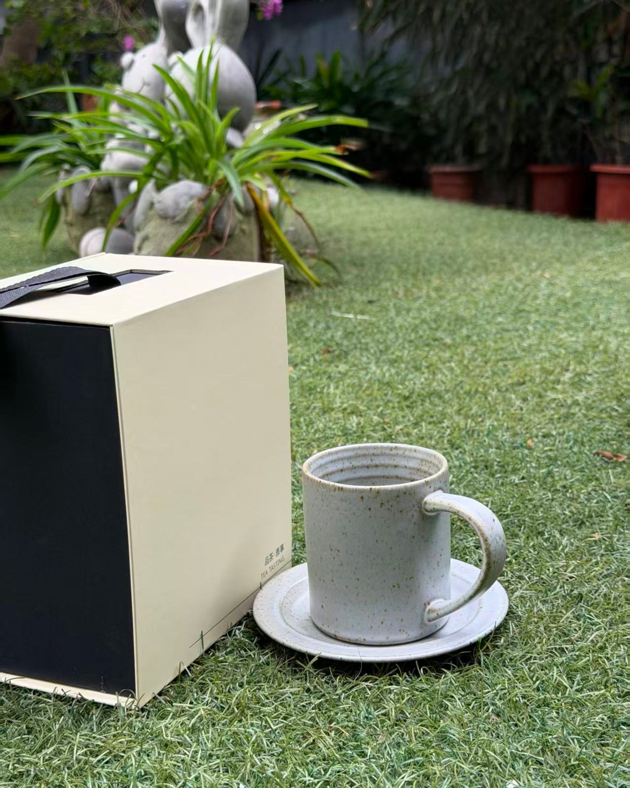 A speckled off-white ceramic mug with a matching saucer sits on green grass next to a cream and black gift box, with a garden background.
