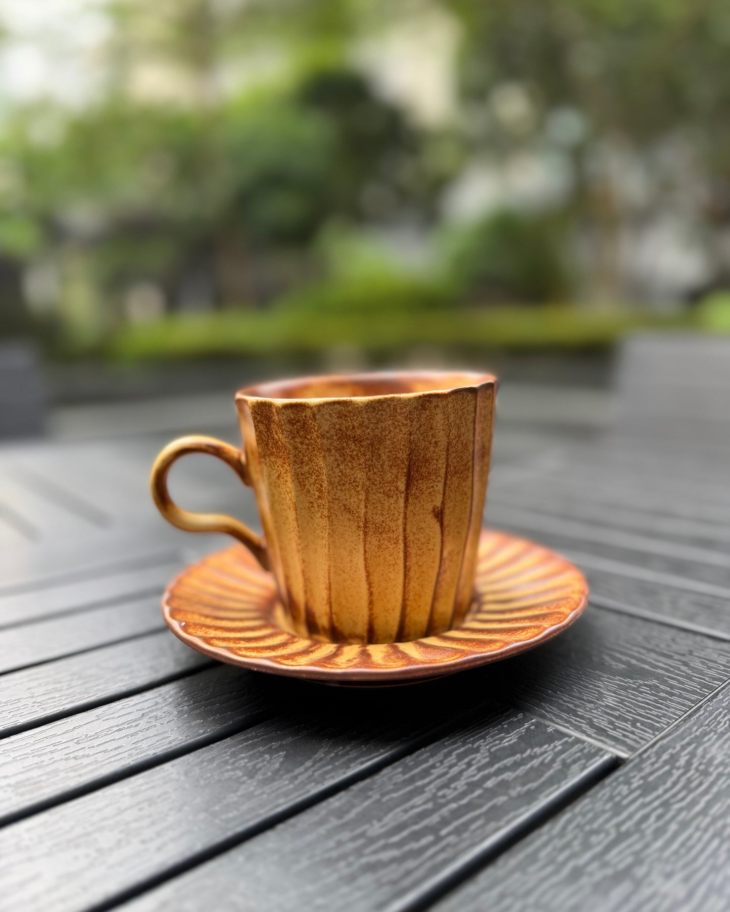 Back view of an amber lines fluted stoneware mug with matching saucer on a dark slatted outdoor table, highlighting the reactive kiln-fired glaze and scalloped saucer edge, with blurred green foliage behind