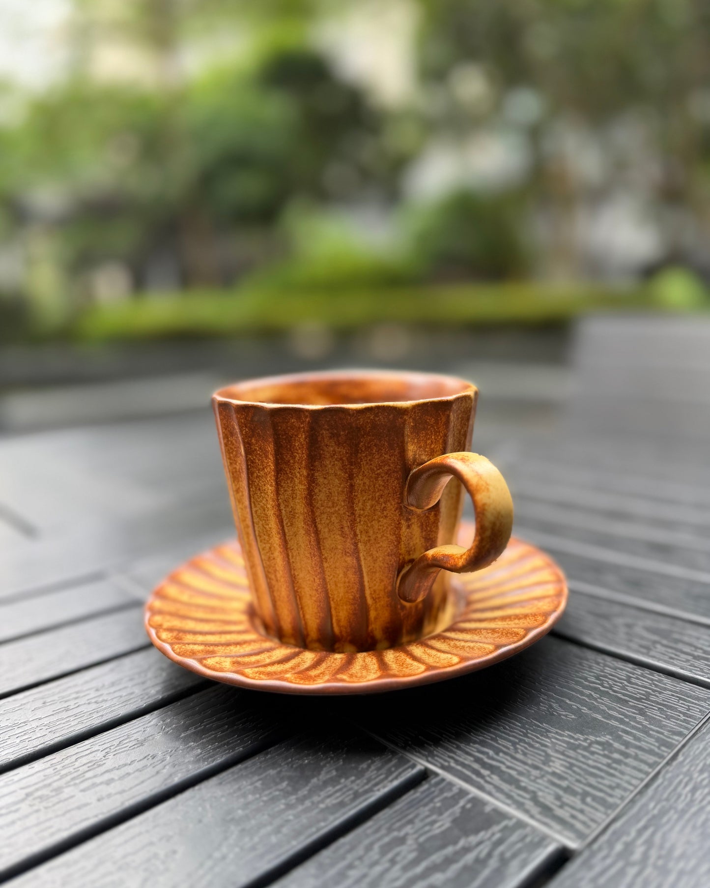 Side view of an amber lines fluted stoneware mug with matching saucer on a dark slatted outdoor table, showing the curved handle detail and warm amber glaze finish, with soft natural light.