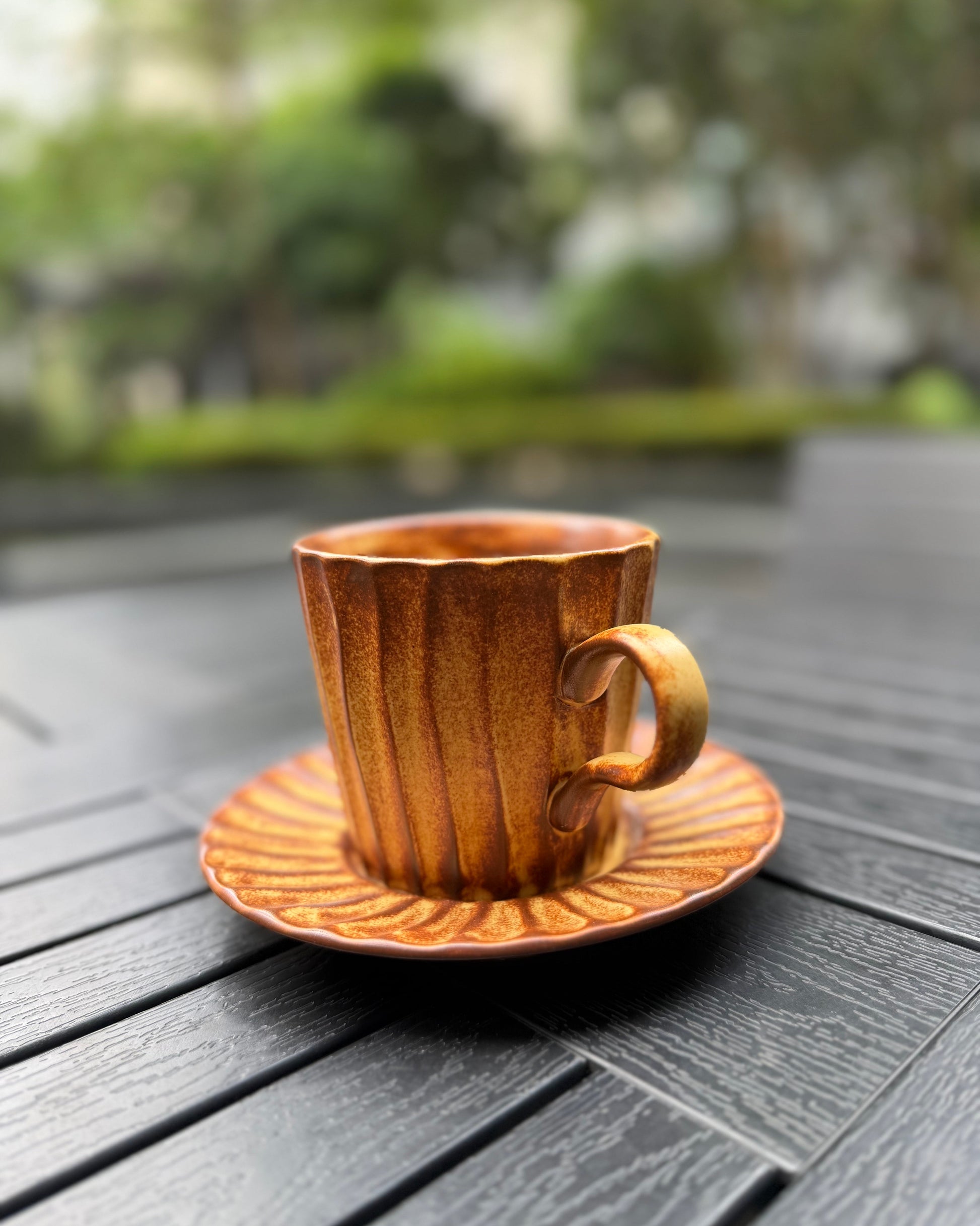 Side view of an amber lines fluted stoneware mug with matching saucer on a dark slatted outdoor table, showing the curved handle detail and warm amber glaze finish, with soft natural light.