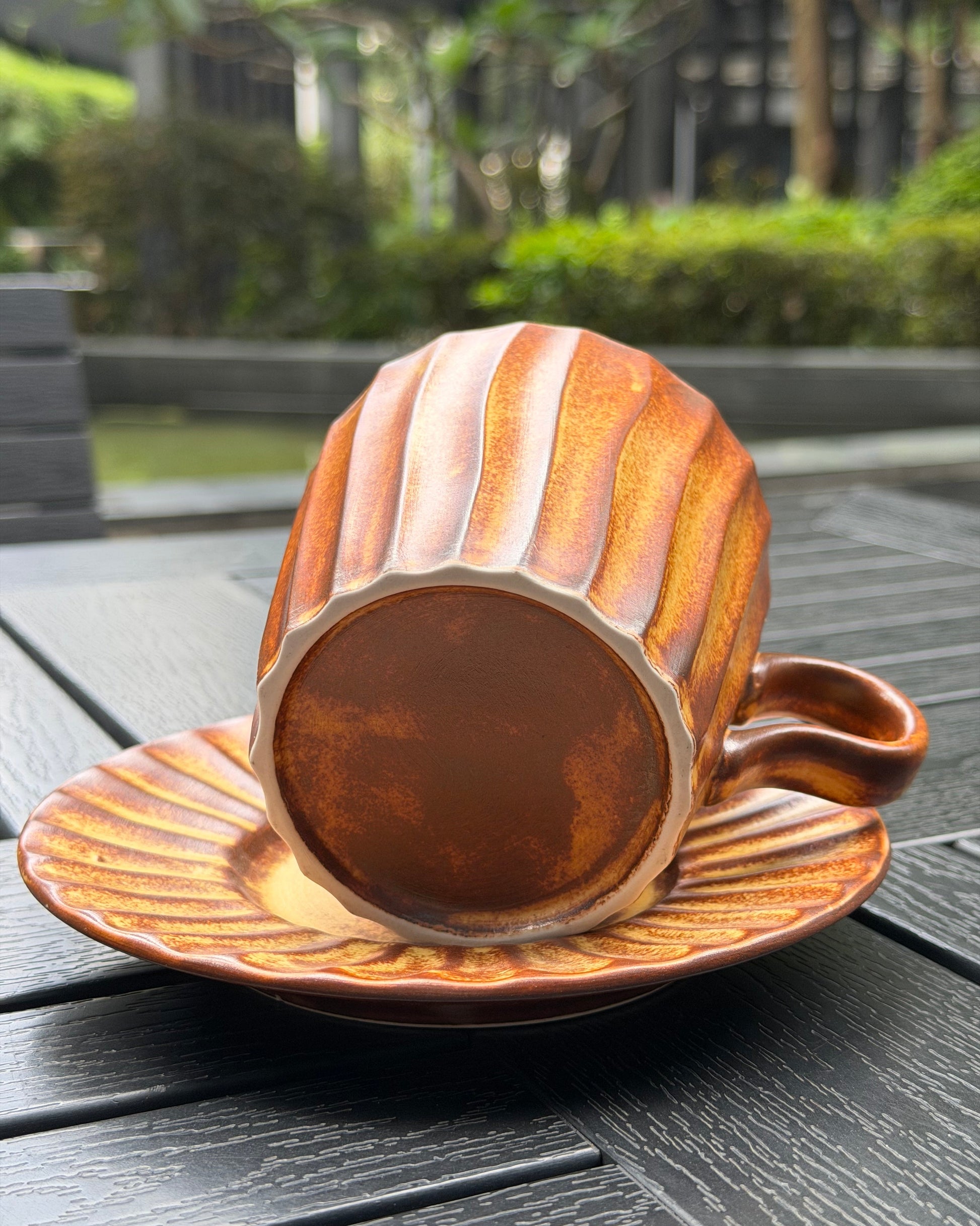 A ribbed ceramic cup and saucer set in a warm terracotta brown color, photographed outdoors on a dark wooden table with a blurred garden background.