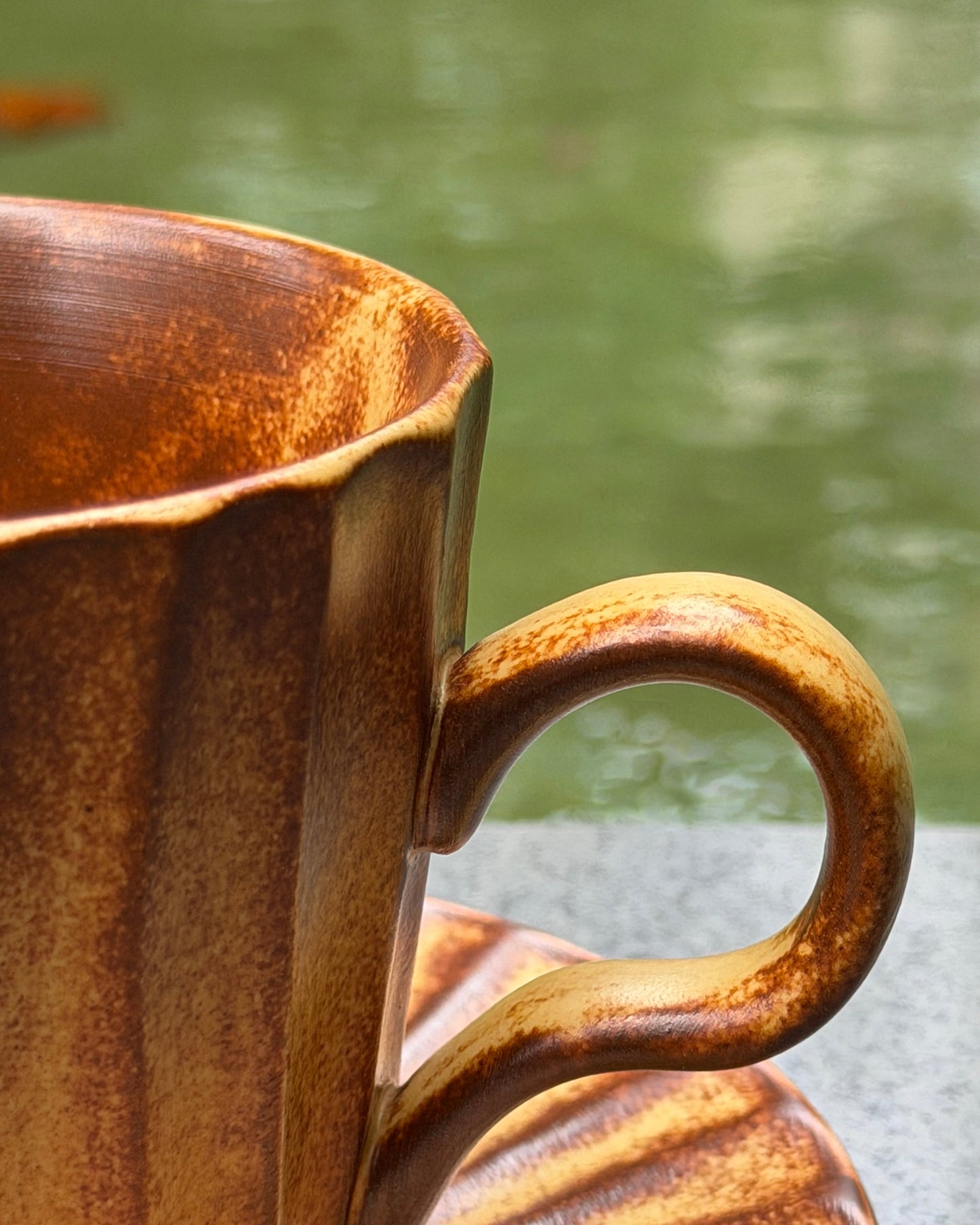 Close-up of a rustic brown ceramic mug with a matching saucer, featuring a textured glaze and a curved handle, set against a blurred green outdoor background.