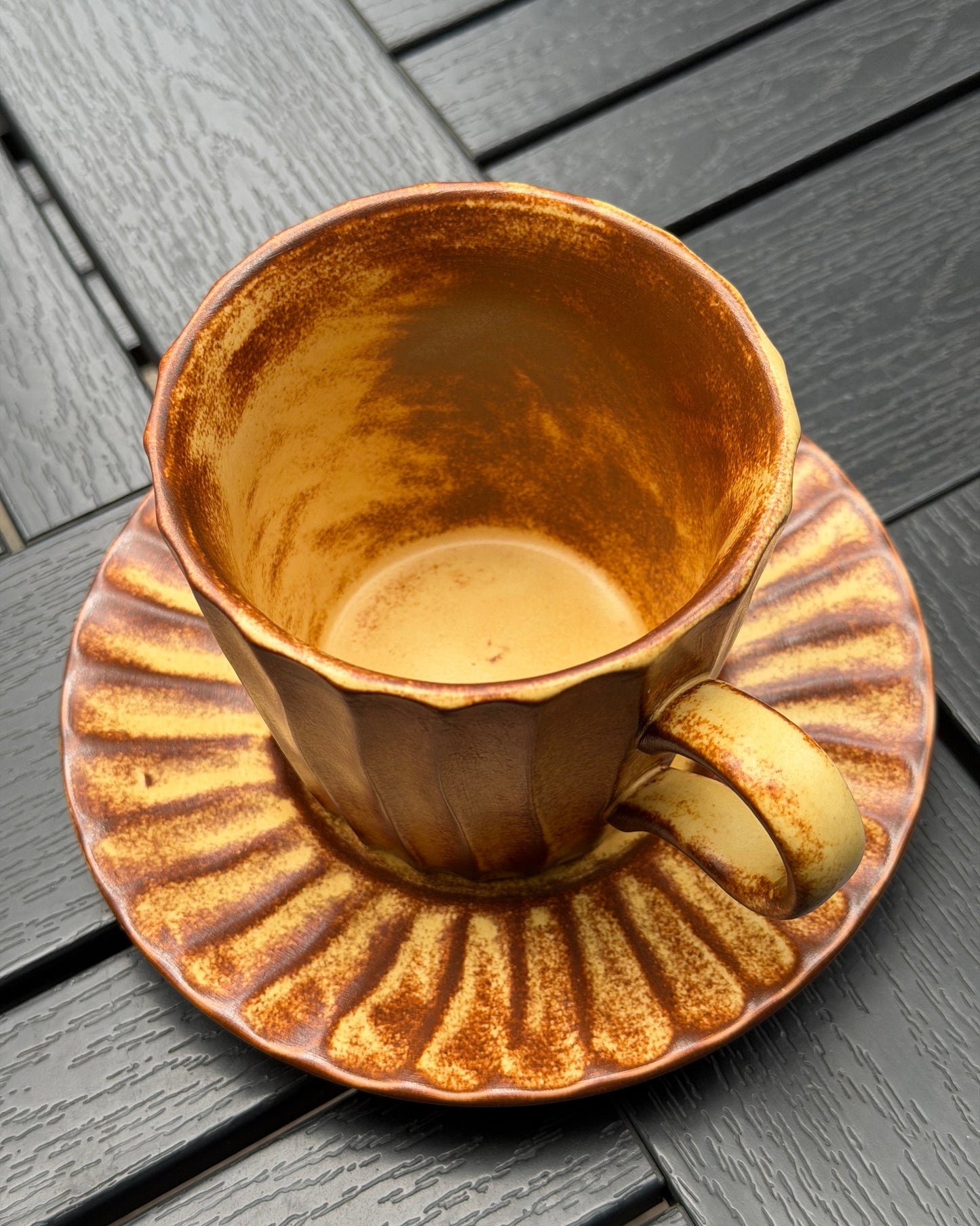A ceramic teacup and saucer set with a metallic copper-brown and gold finish, shown on a dark gray slatted wooden surface.