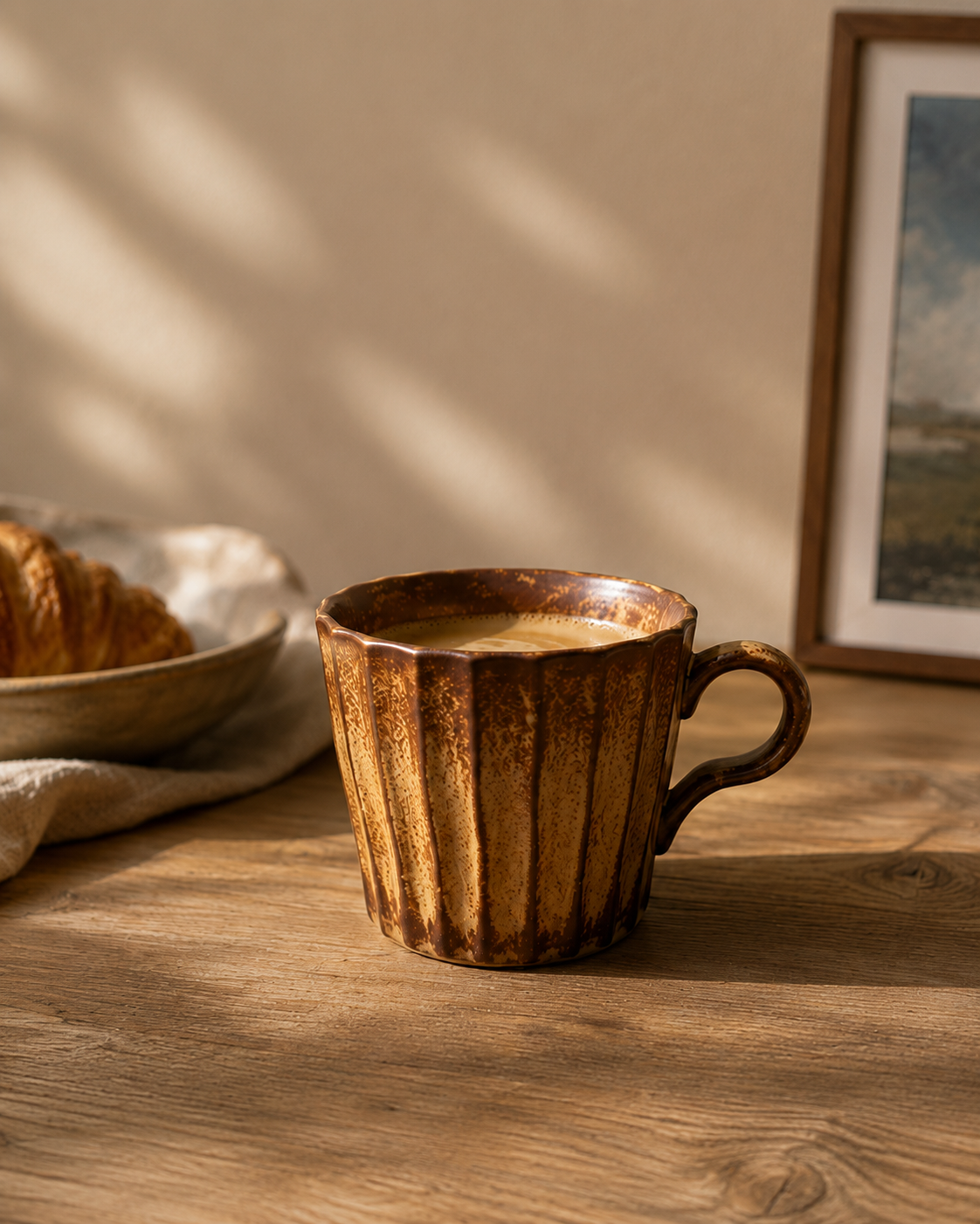 A rustic brown ceramic mug with a ribbed fluted design sits on a wooden table, filled with coffee and set in a warm, sunlit room with a blurred croissant and framed picture in the background.