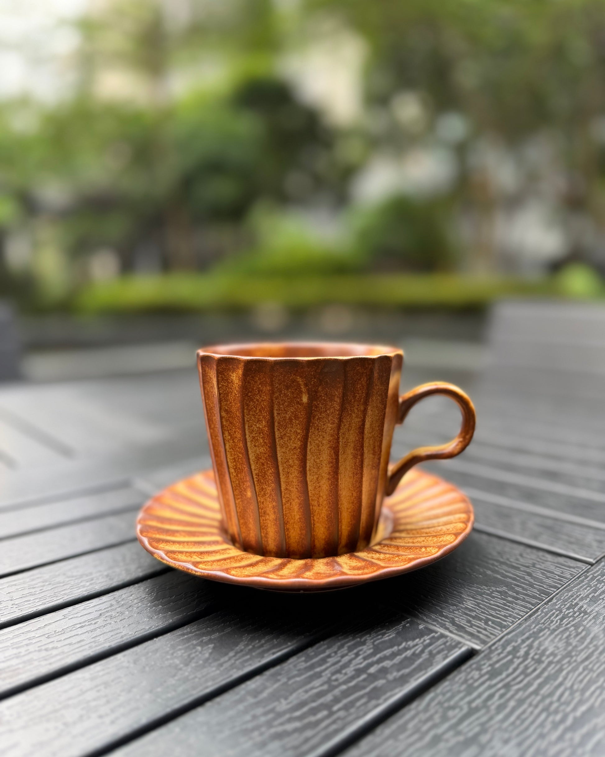 Amber lines fluted stoneware mug with matching saucer on a dark slatted outdoor table, showcasing the vertical ribbed texture and warm amber glaze, with blurred green foliage in the background