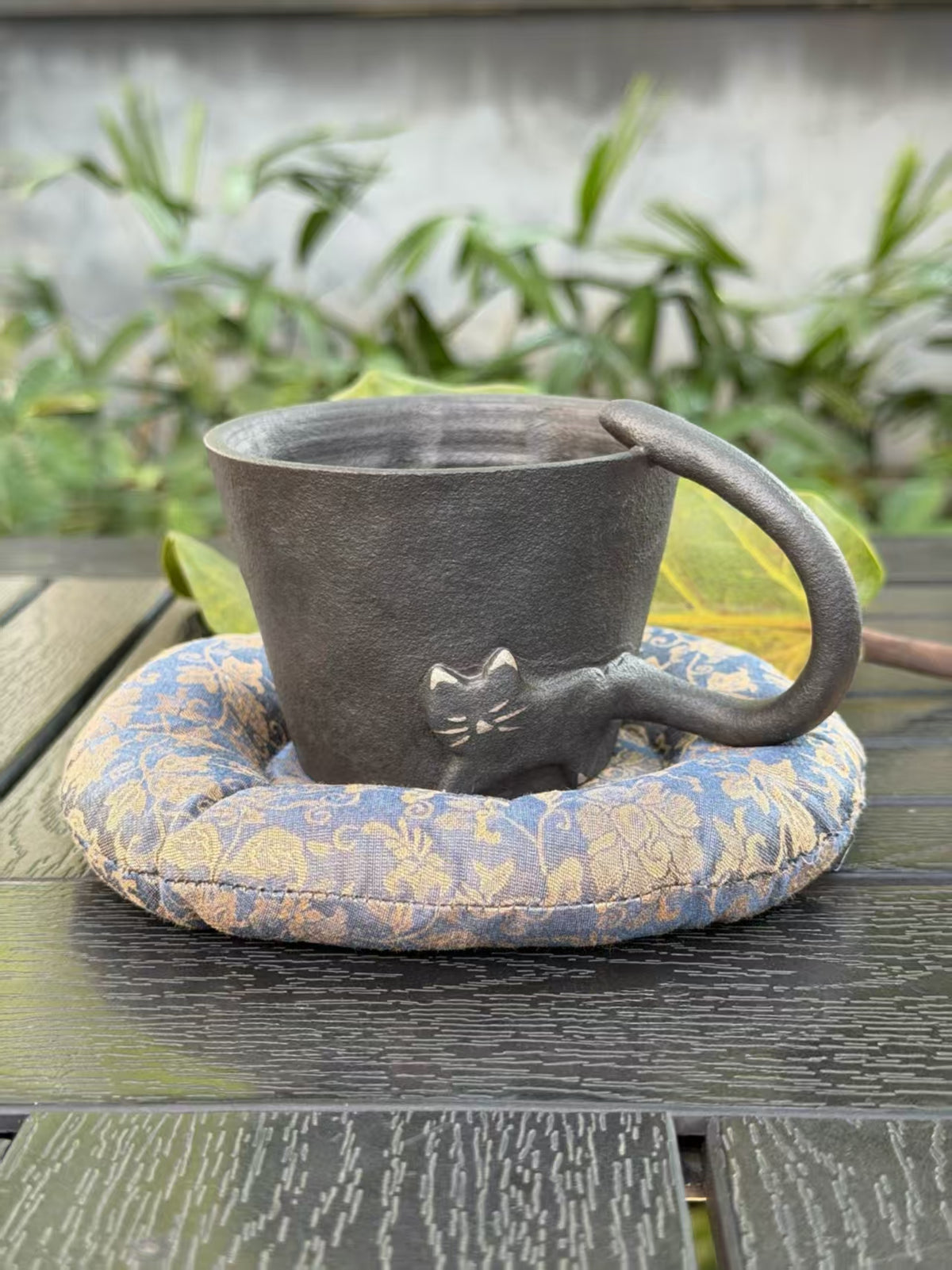 Front view of black cat ceramic cup with sculptural tail handle and blue and gold patterned cushion