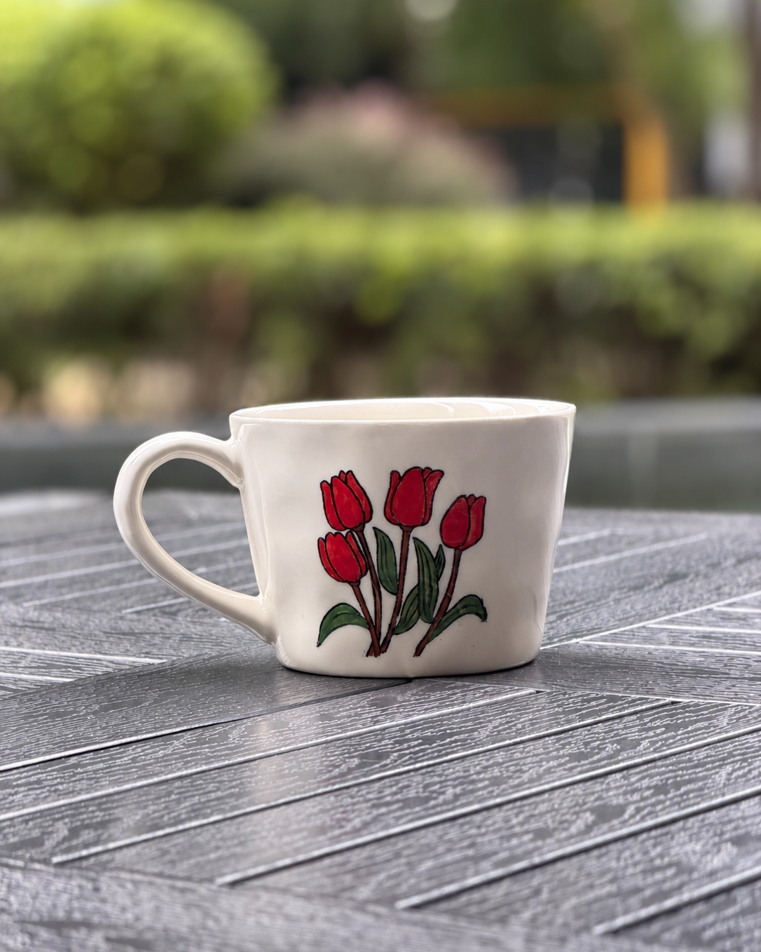 A white ceramic coffee mug with a red tulip and green leaf design, sitting on a gray textured outdoor table with a blurred garden background.