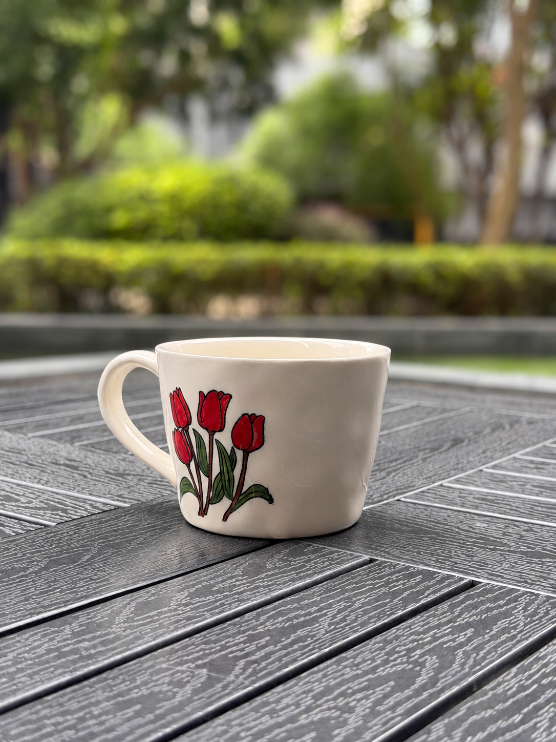 Side-angle view of a cream ceramic coffee mug with hand-painted red tulips against a blurred garden