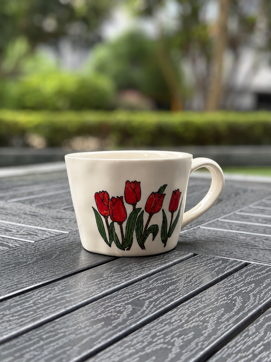 Angled side view of a cream ceramic coffee mug with a hand-painted red tulip garden design and curved handle