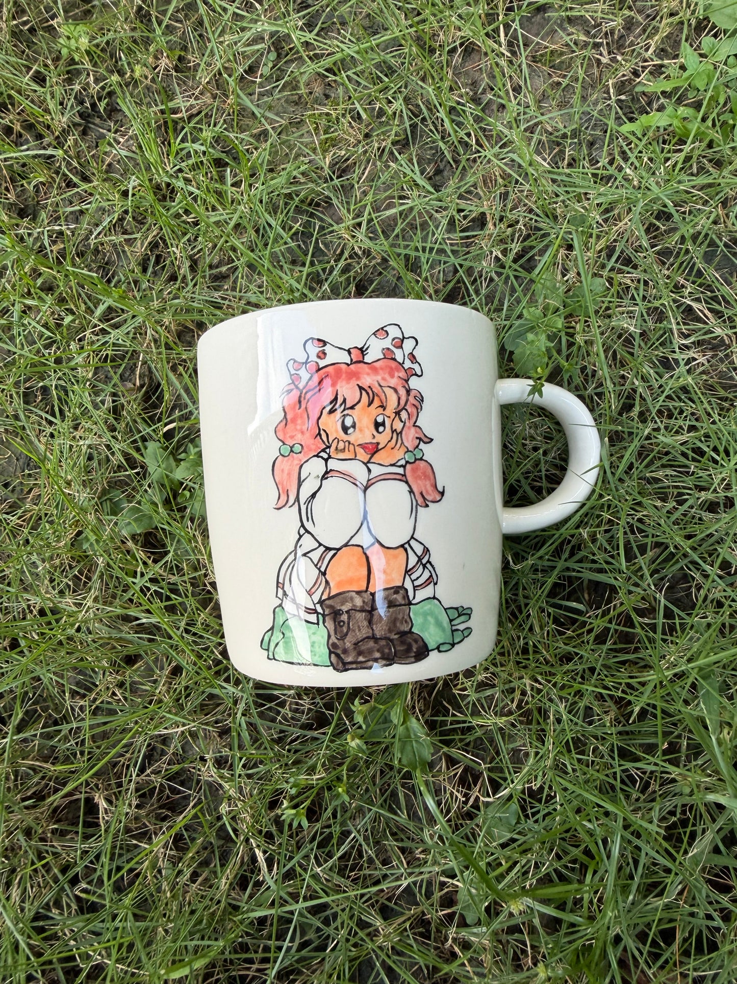 Hand-painted ceramic mug lying on green grass, front side showing full-body seated girl with pink pigtails, white dress, and brown boots