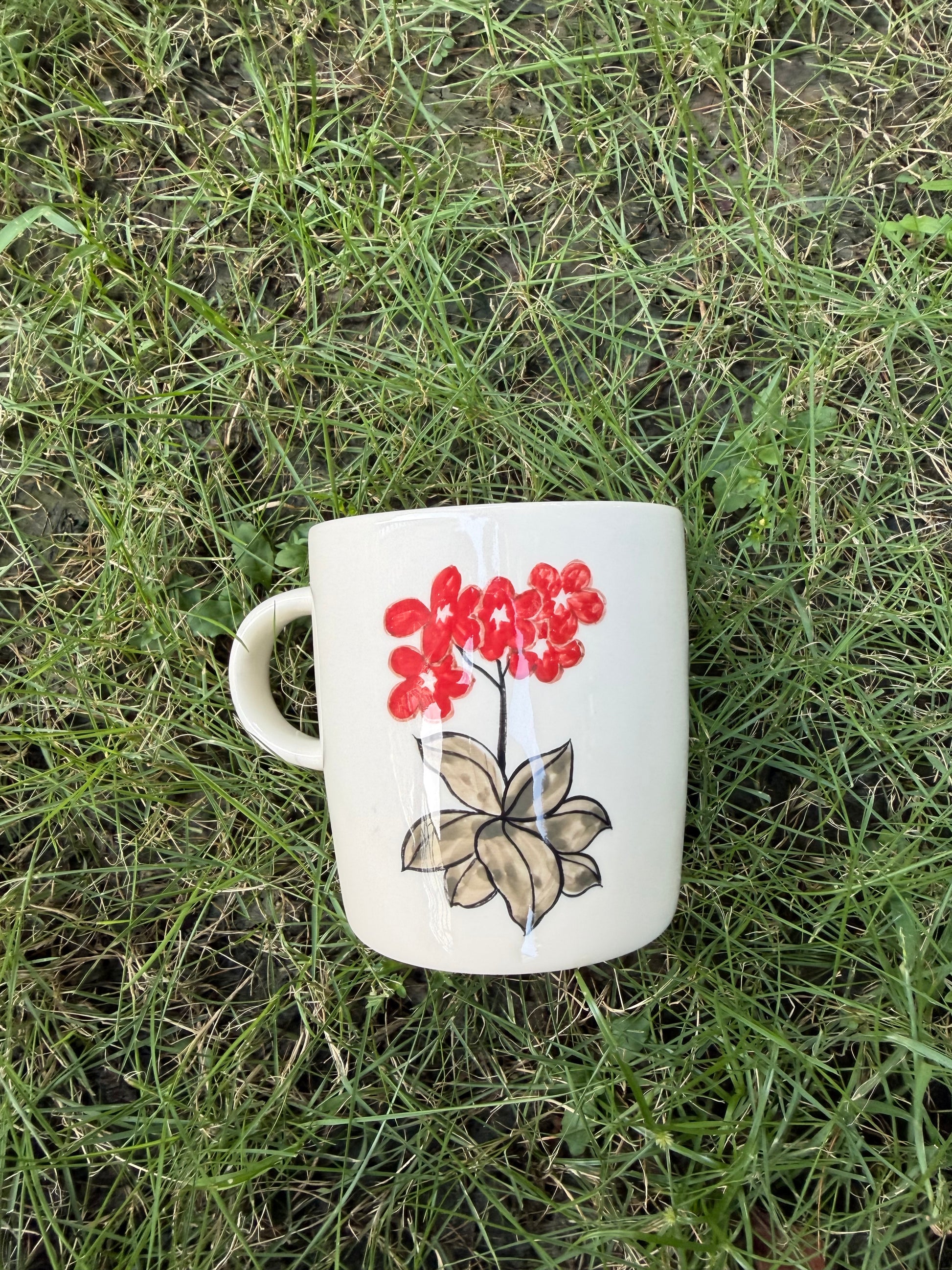 Hand-painted ceramic mug lying on green grass, back side showing vivid red floral cluster with loose ink-style brown leaves