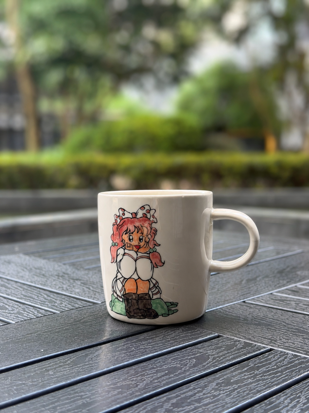 A white ceramic coffee mug with a colorful anime-style character illustration, sitting on a dark gray slatted outdoor table with a blurred green garden background.