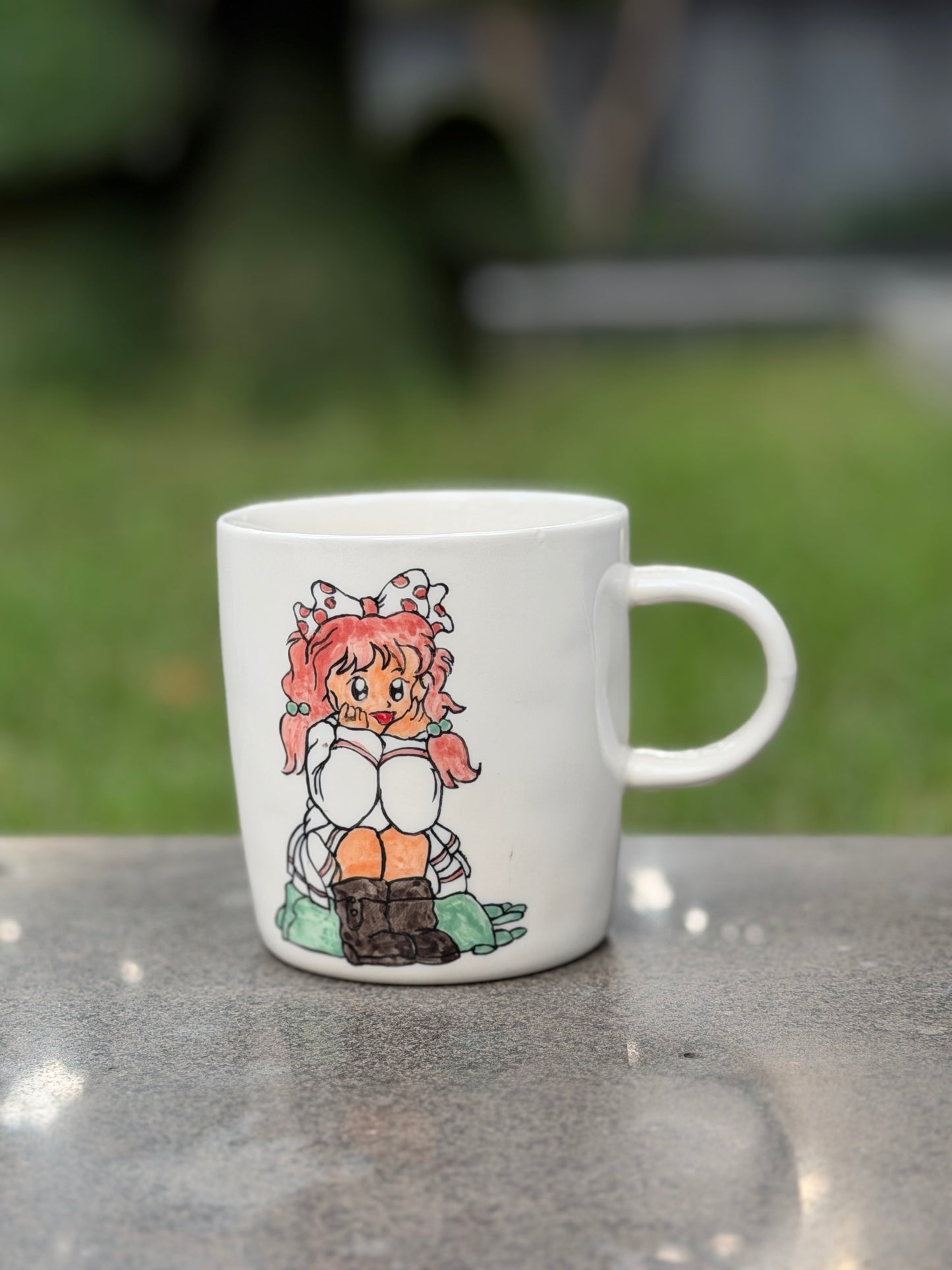 Hand-painted ceramic mug on a grey stone surface outdoors, front side showing seated girl with pink pigtails and polka-dot bow, green garden background