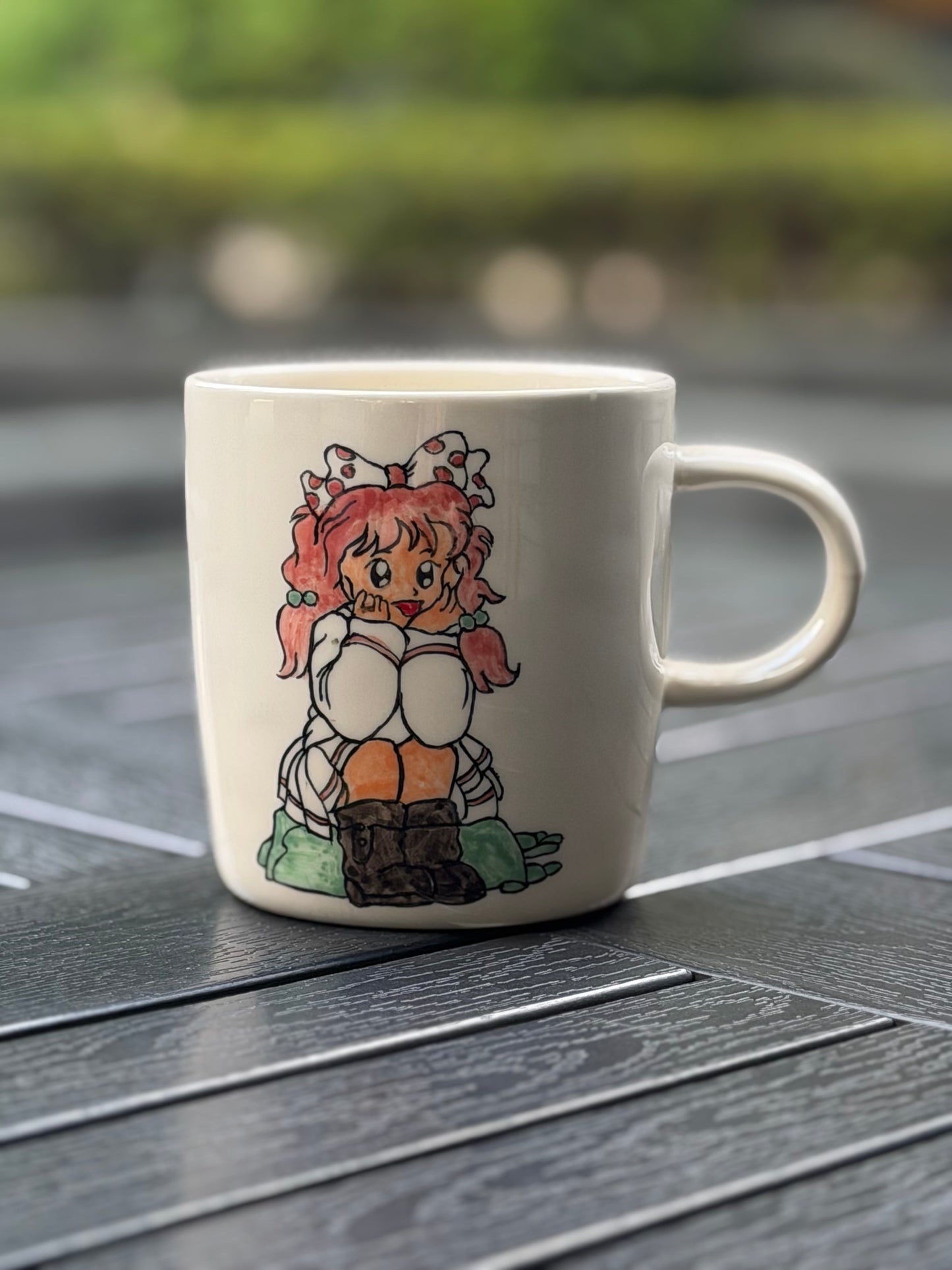 Hand-painted ceramic mug on a dark wood outdoor table, front side showing seated girl with pink pigtails and polka-dot bow