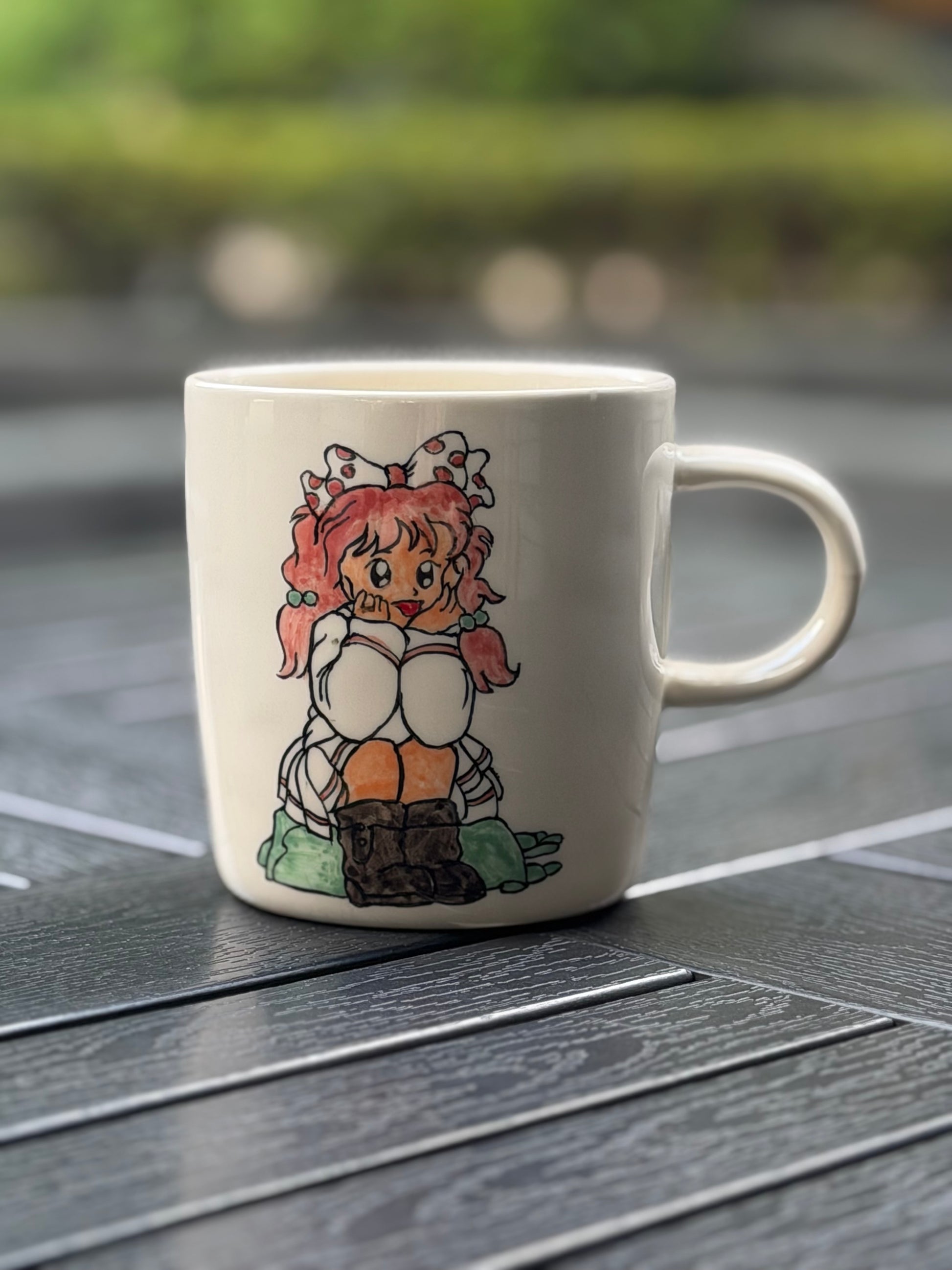 Hand-painted ceramic mug on a dark wood outdoor table, front side showing seated girl with pink pigtails and polka-dot bow