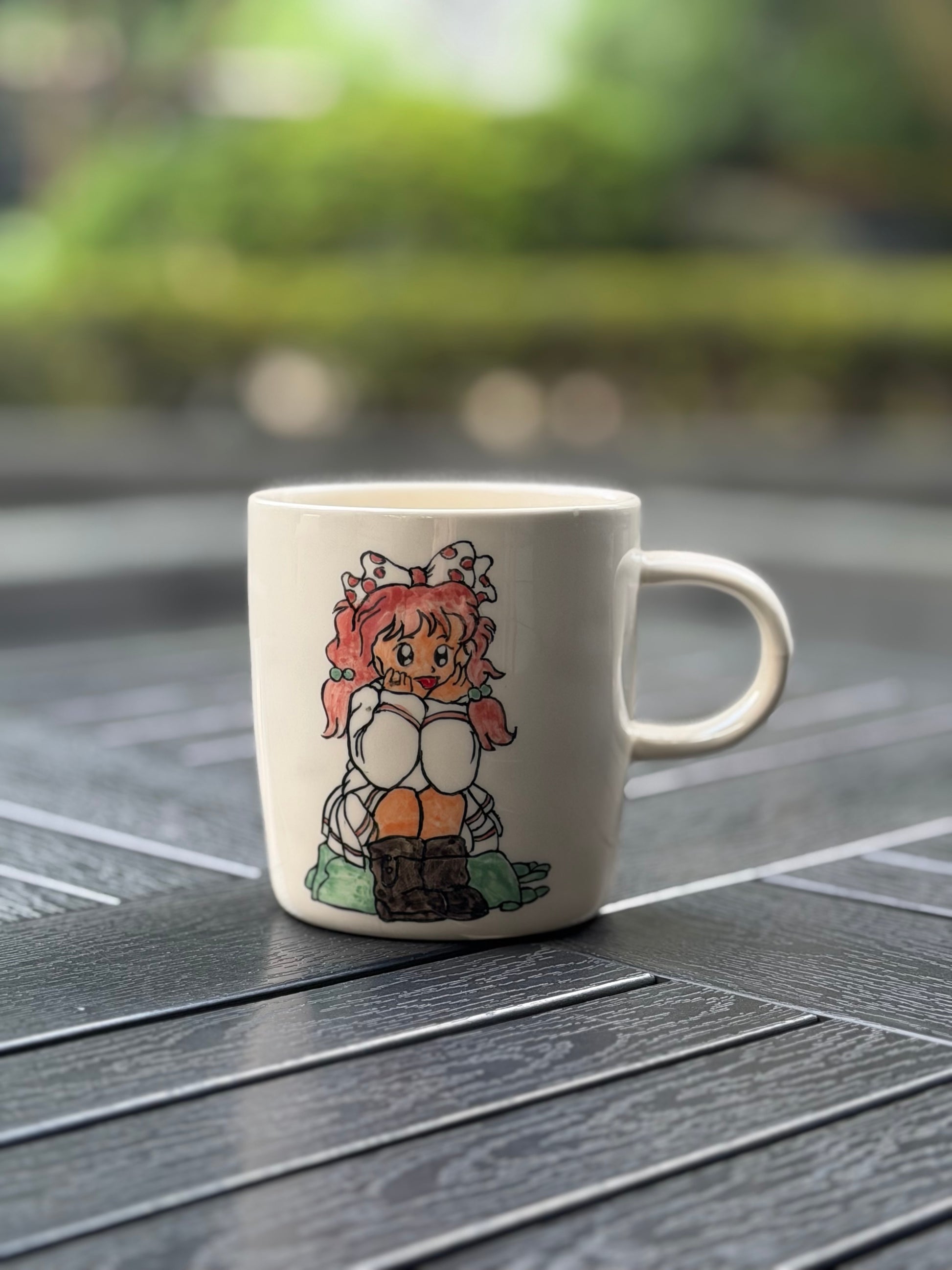 Hand-painted ceramic mug on a dark wood outdoor table, front side showing a seated girl with pink pigtails, polka-dot bow, white dress, and brown boots