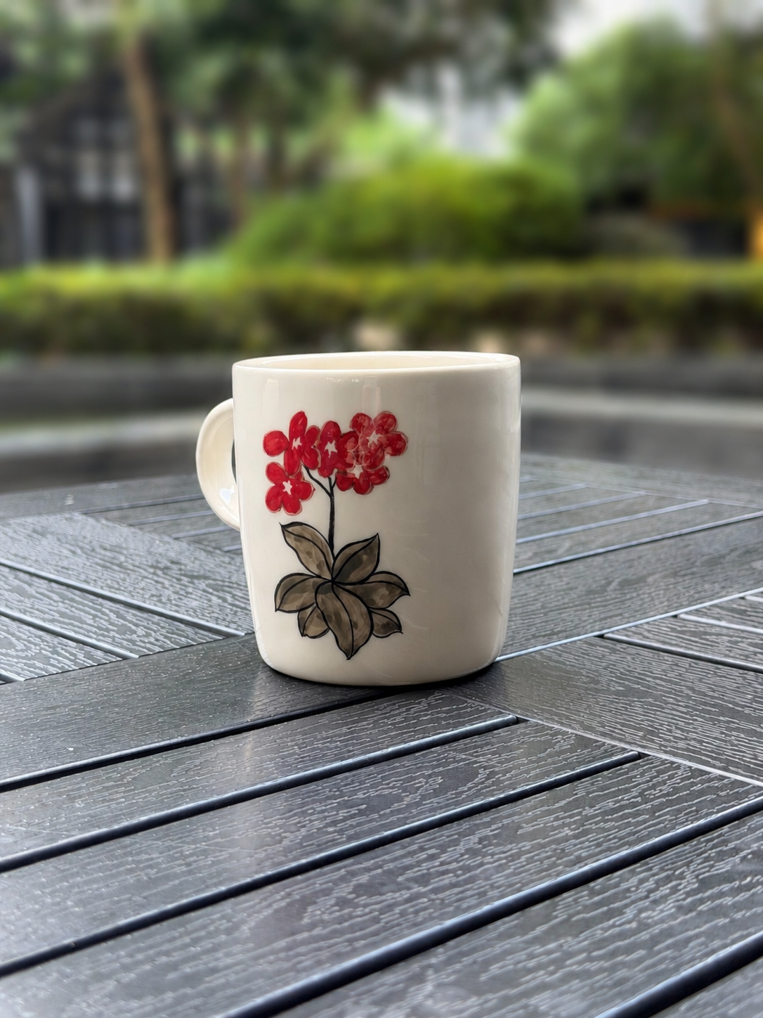 Hand-painted ceramic mug on a dark wood outdoor table, back side showing red floral cluster with brown ink leaves, viewed from front-right angle