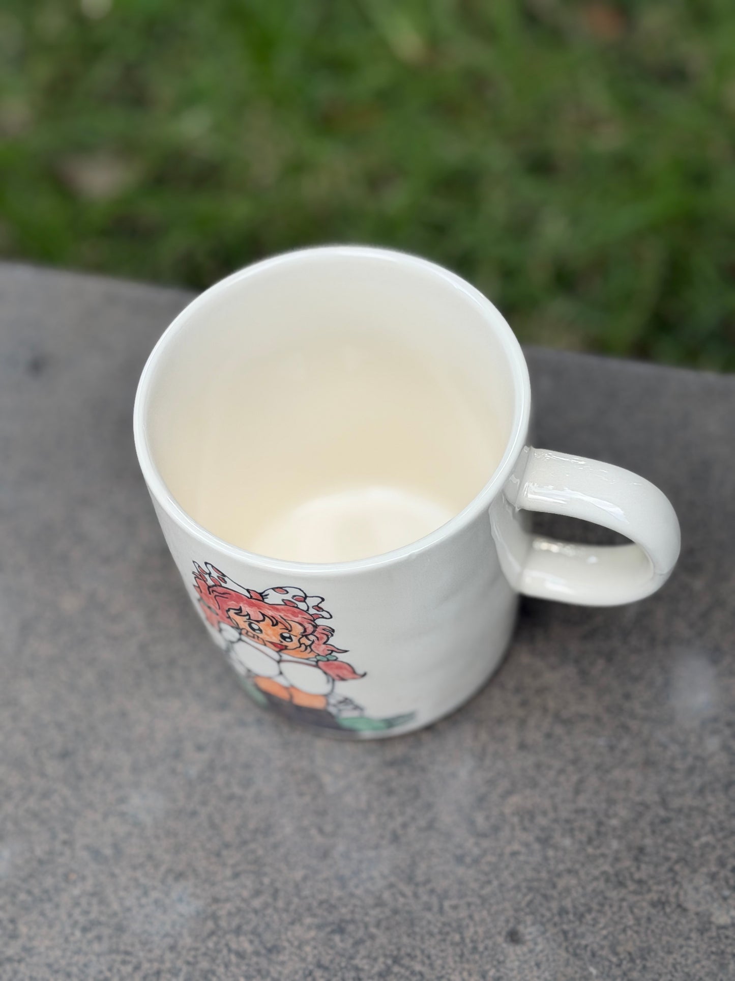 Angled top view of white ceramic mug on an outdoor surface, showing smooth cream-glazed interior and rounded handle