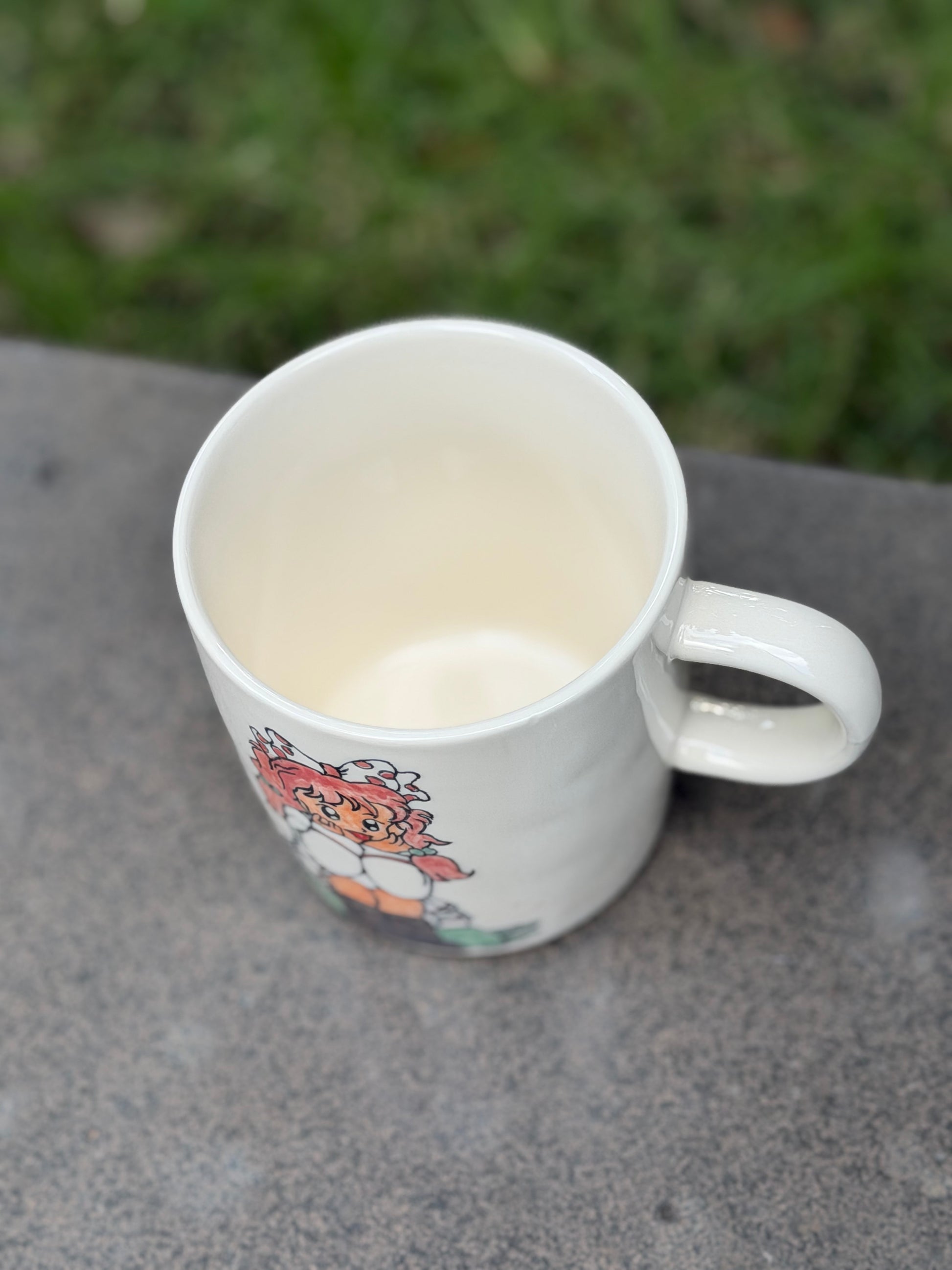 Angled top view of white ceramic mug on an outdoor surface, showing smooth cream-glazed interior and rounded handle