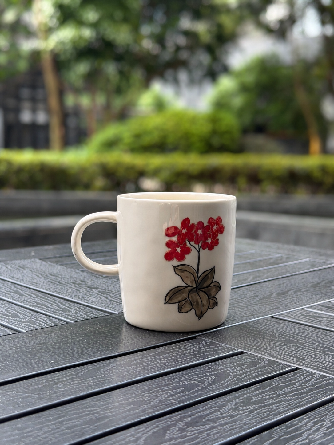Hand-painted ceramic mug on a dark wood outdoor table, back side showing red floral cluster with earthy brown ink leaves