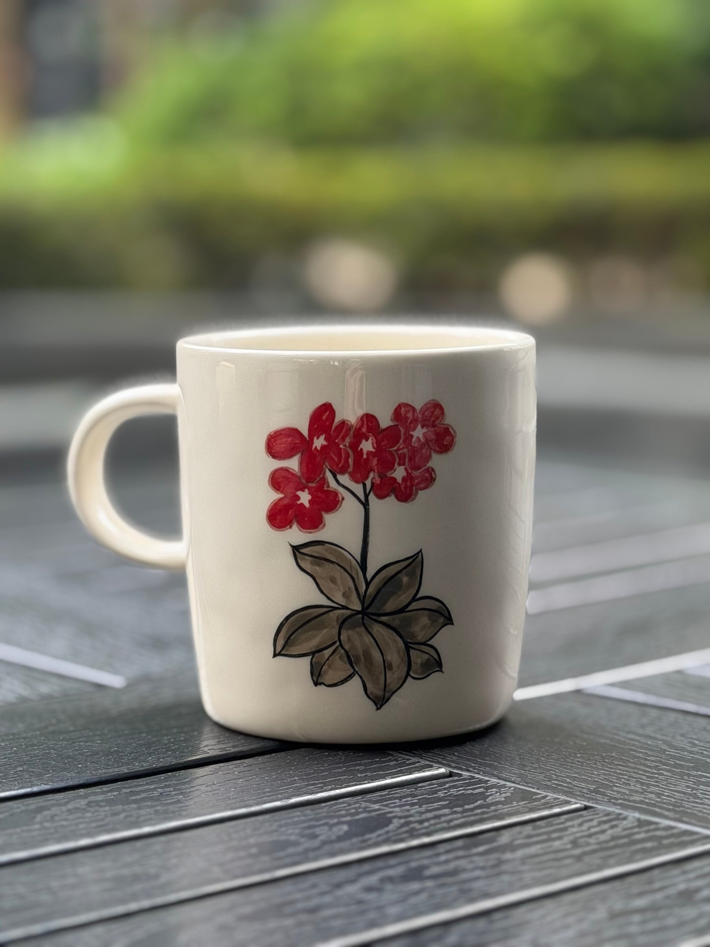 Hand-painted ceramic mug on a dark grey lattice outdoor table, back side showing a vivid red flower cluster with grey-brown ink leaves, blurred green garden background