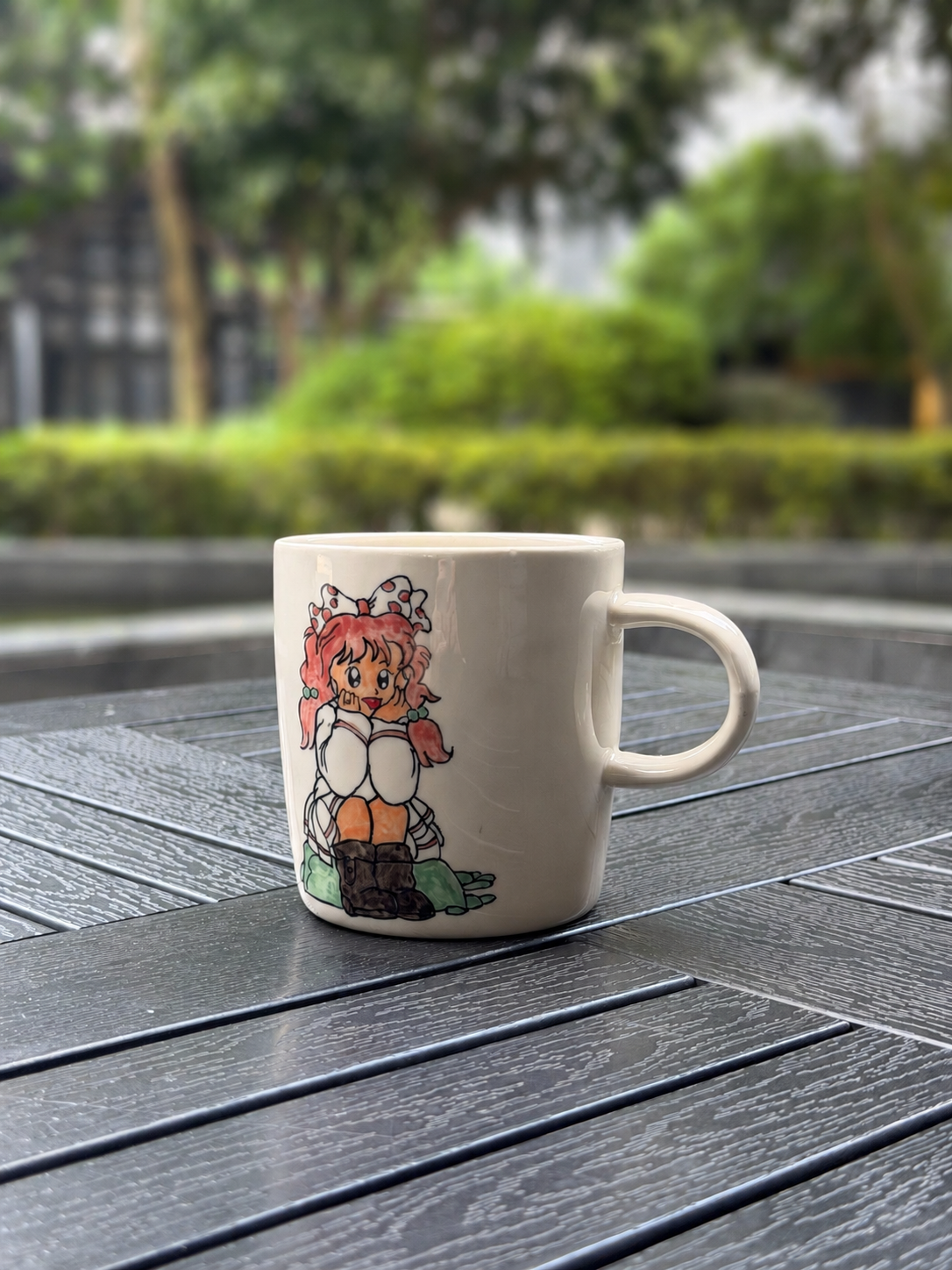 Hand-painted ceramic mug on a dark wood outdoor table, front side showing seated girl with pink pigtails, viewed from front-right angle