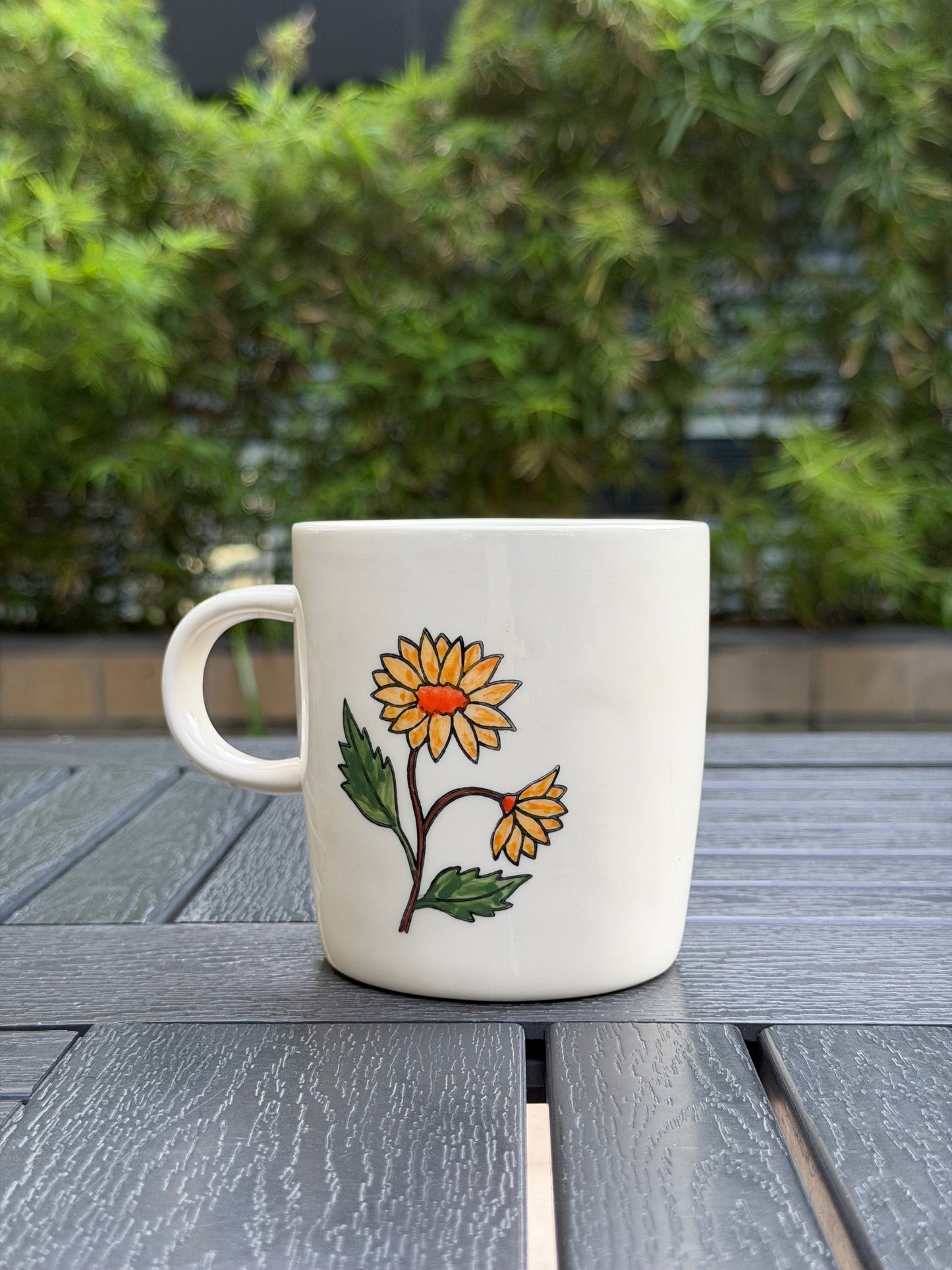 Hand-painted golden chrysanthemum ceramic mug, rear angled view — one full bloom and one side-facing flower, handle on left, green foliage backdrop