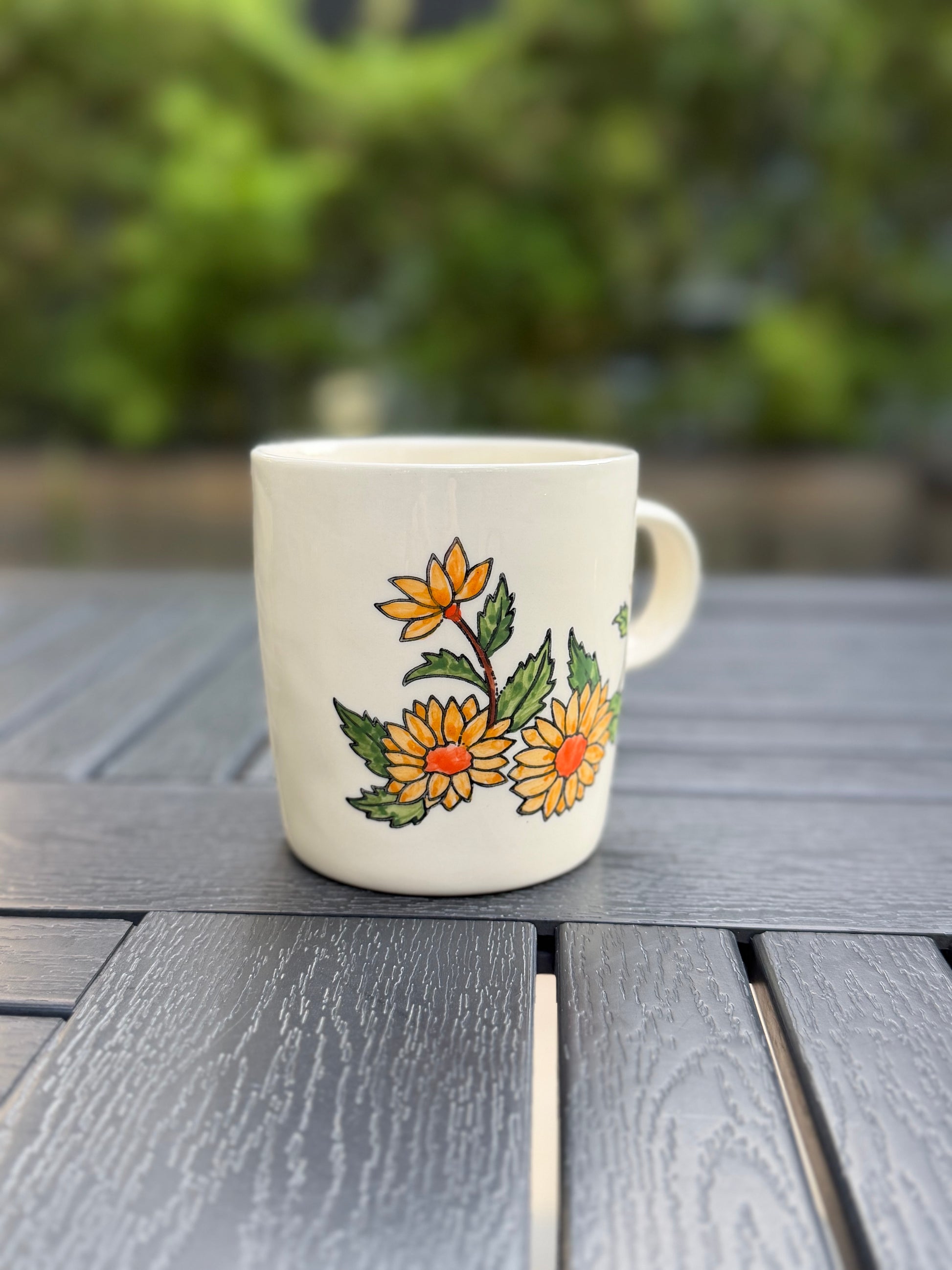 Hand-painted golden chrysanthemum ceramic mug, side view — one bud and two full blooms with orange centers and green leaves on white stoneware, soft green garden backdrop