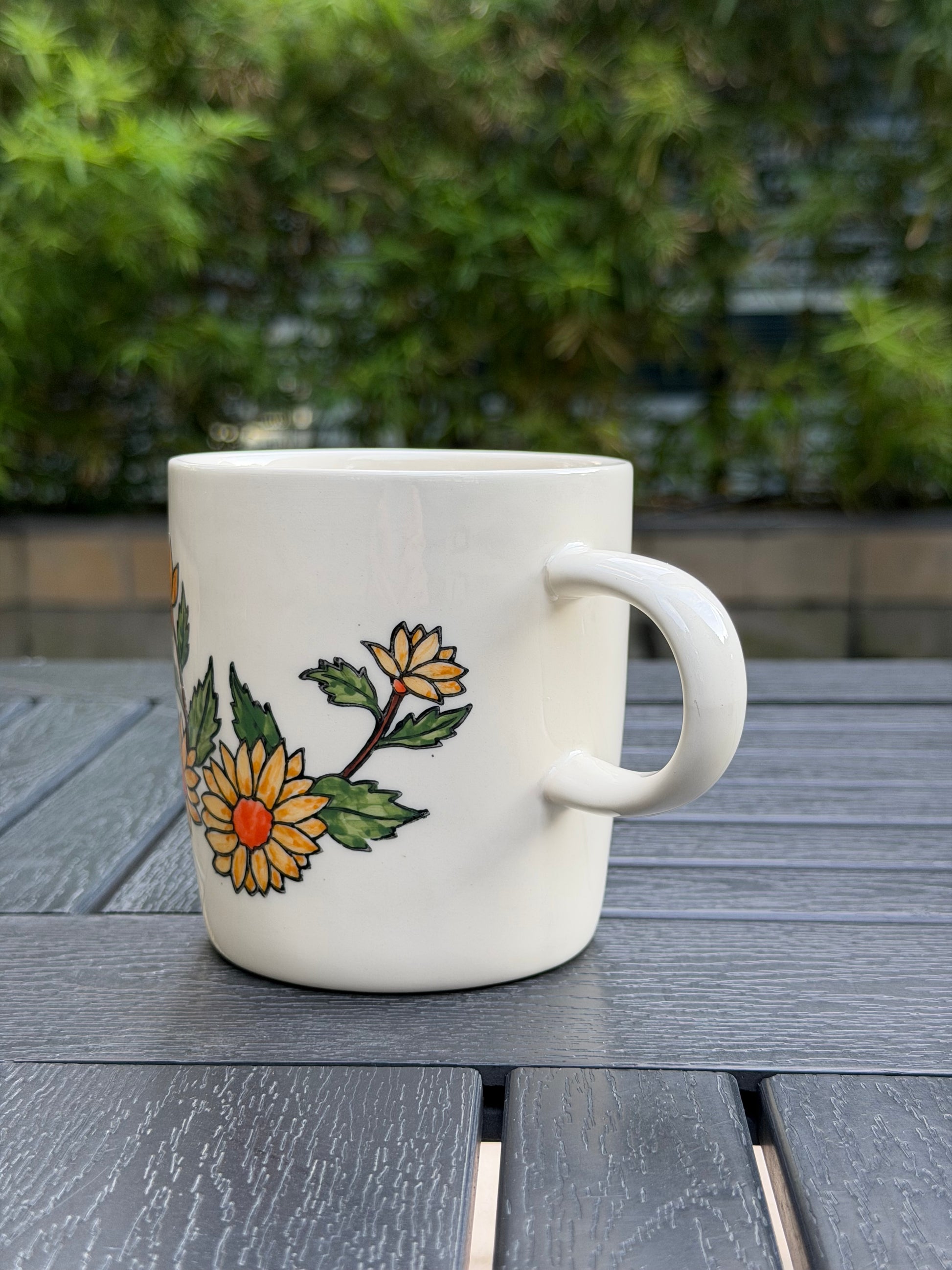 Hand-painted golden chrysanthemum ceramic mug, right-handle side view — two blooms with orange centers and budding flower, white stoneware on outdoor wood table