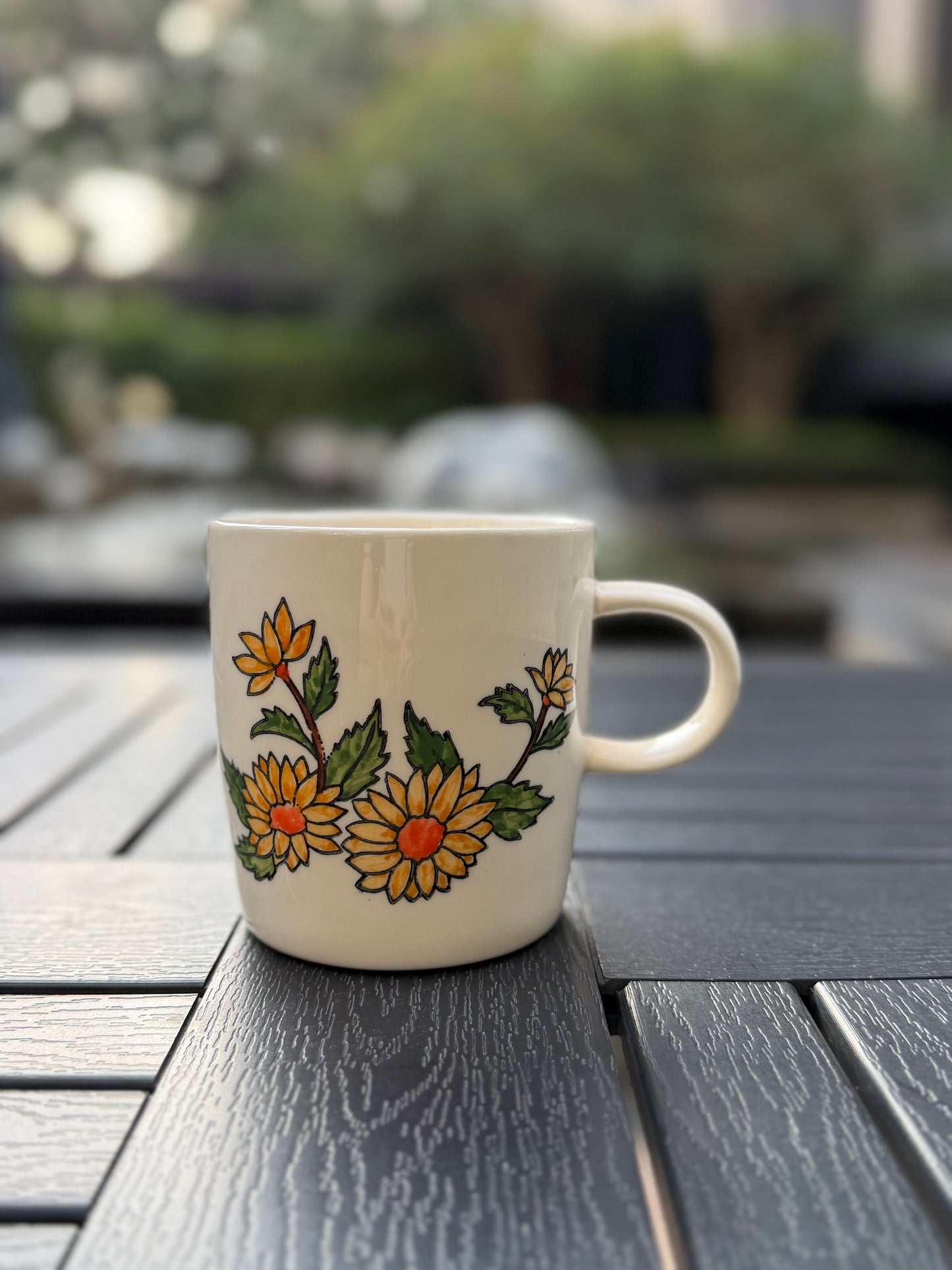 Hand-painted golden chrysanthemum ceramic mug, front view — yellow blooms with orange centers on white stoneware, handle on right, dark foliage backdrop