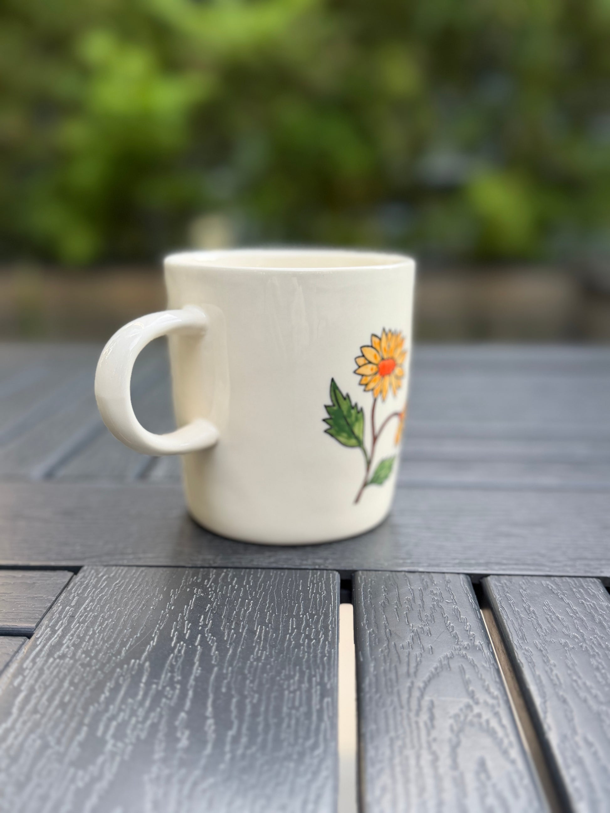 Hand-painted golden chrysanthemum ceramic mug, handle-left side view — partial bloom and bud visible on right edge, white stoneware, low-angle shot on outdoor wood table with soft green backdrop