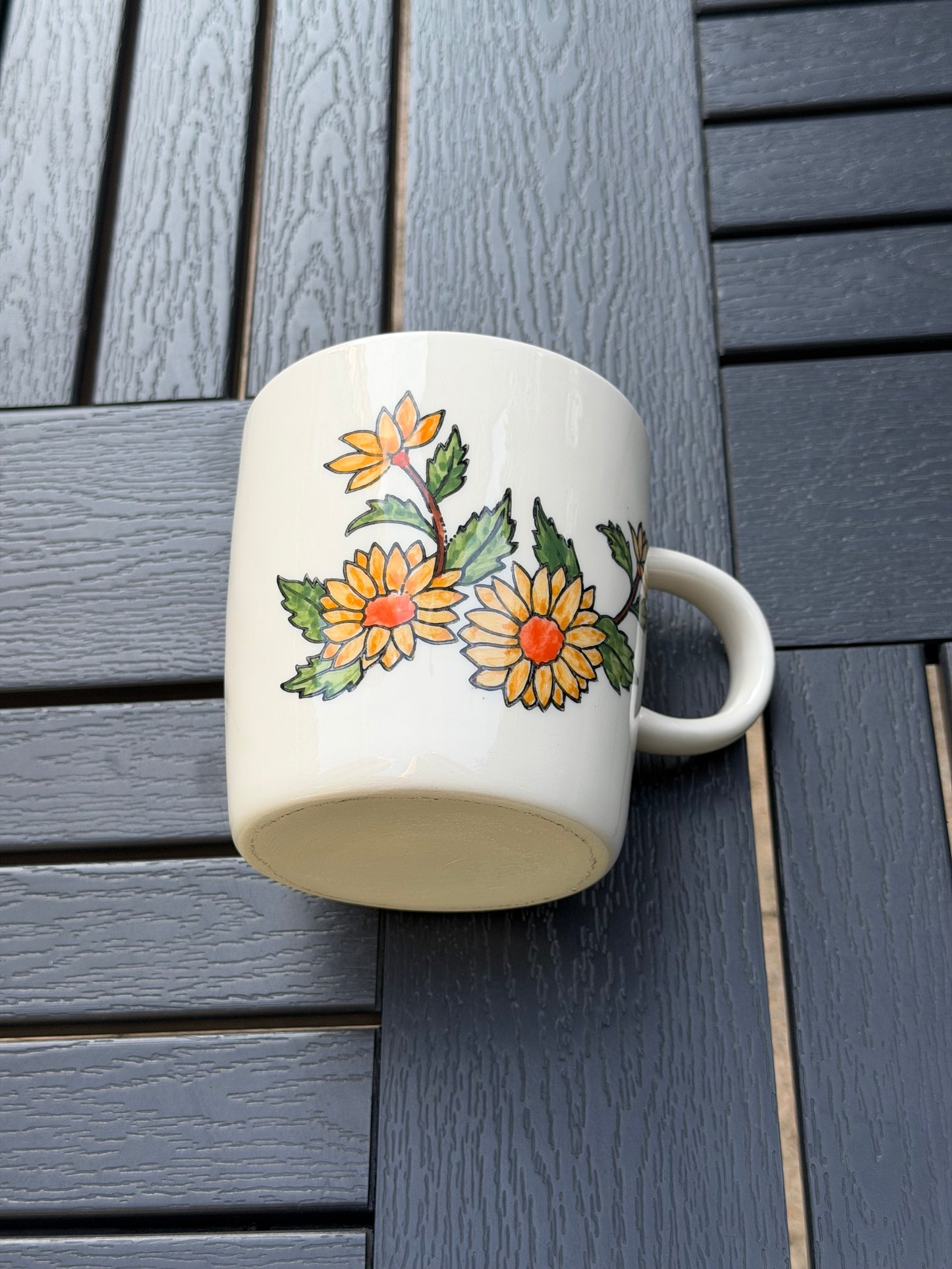 Golden chrysanthemum ceramic mug lying on its side — hand-painted yellow blooms with orange centers and green leaves visible on white stoneware, overhead view