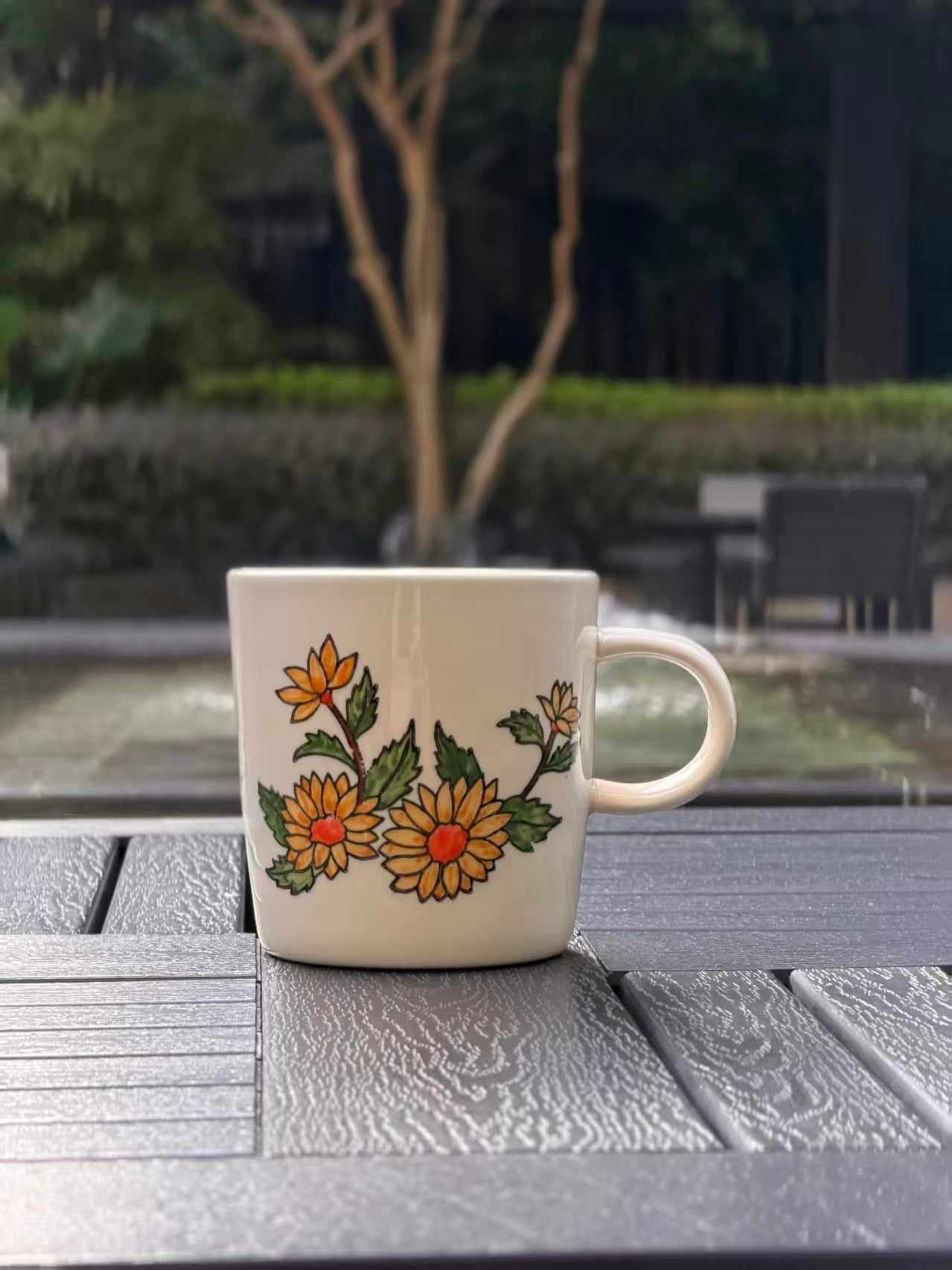 Hand-painted golden chrysanthemum ceramic mug, front view — yellow blooms with orange centers, white stoneware, styled on outdoor table with urban garden backdrop