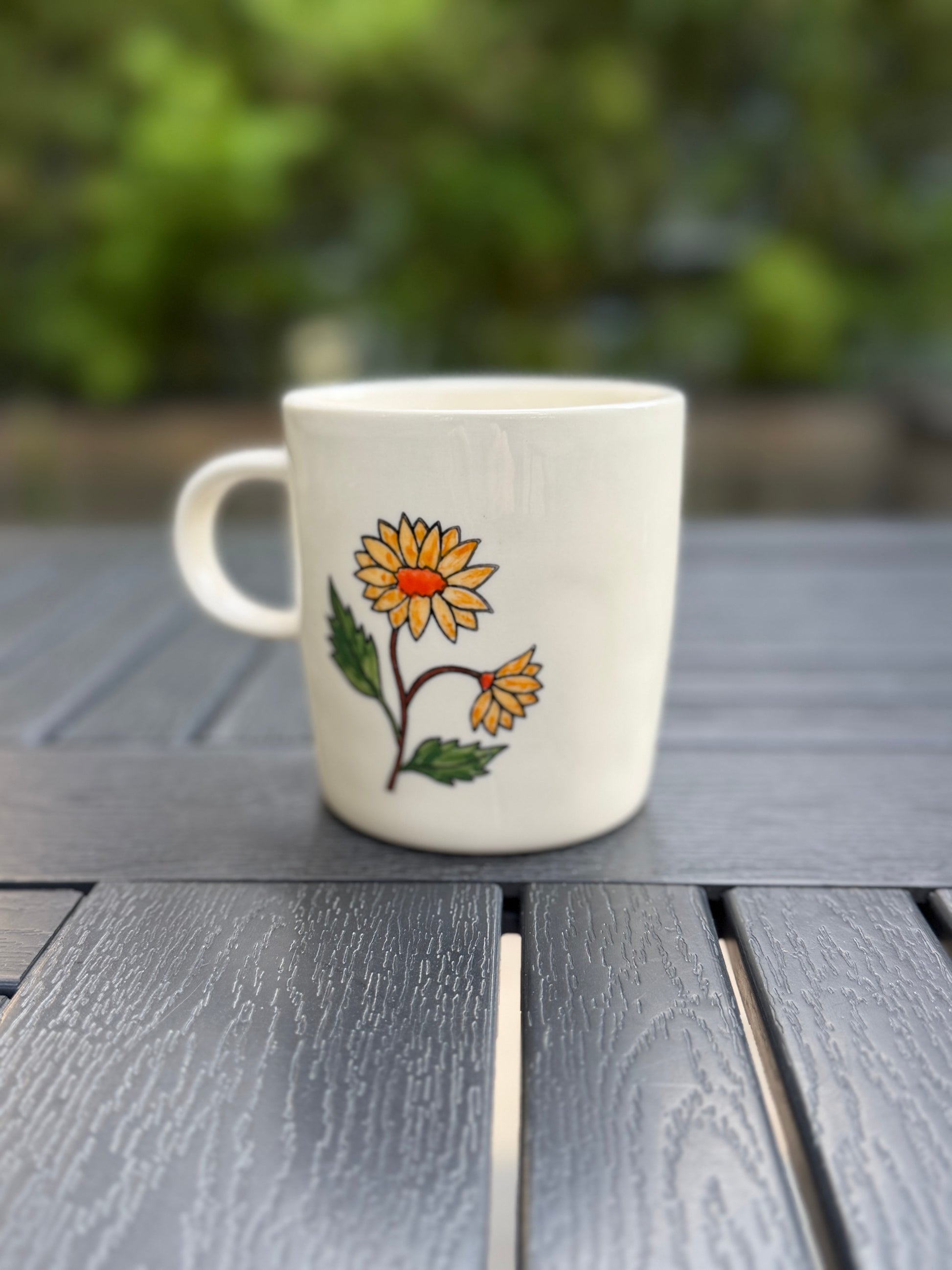Hand-painted golden chrysanthemum ceramic mug, back view — one full bloom and one bud, handle on left, green garden backdrop