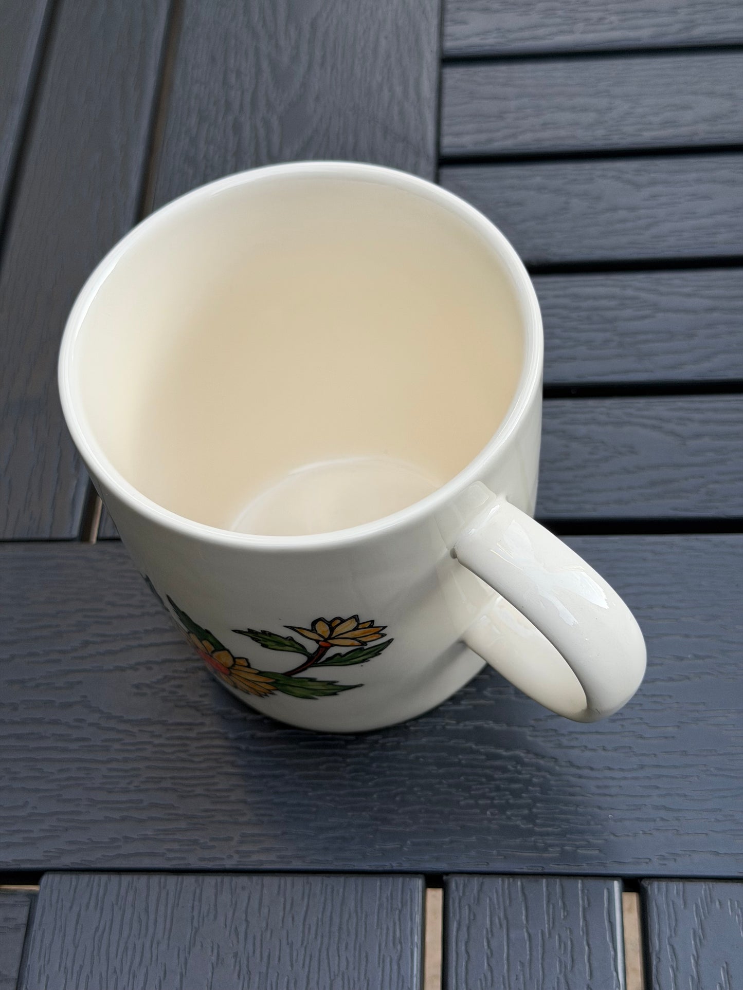 Top-down view of hand-painted golden chrysanthemum ceramic mug — cream-glazed interior, handle on right, flower detail visible at base on outdoor wood table
