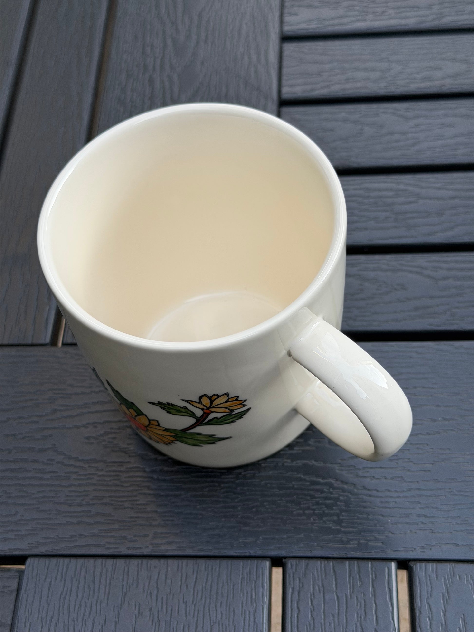 Top-down view of hand-painted golden chrysanthemum ceramic mug — cream-glazed interior, handle on right, flower detail visible at base on outdoor wood table