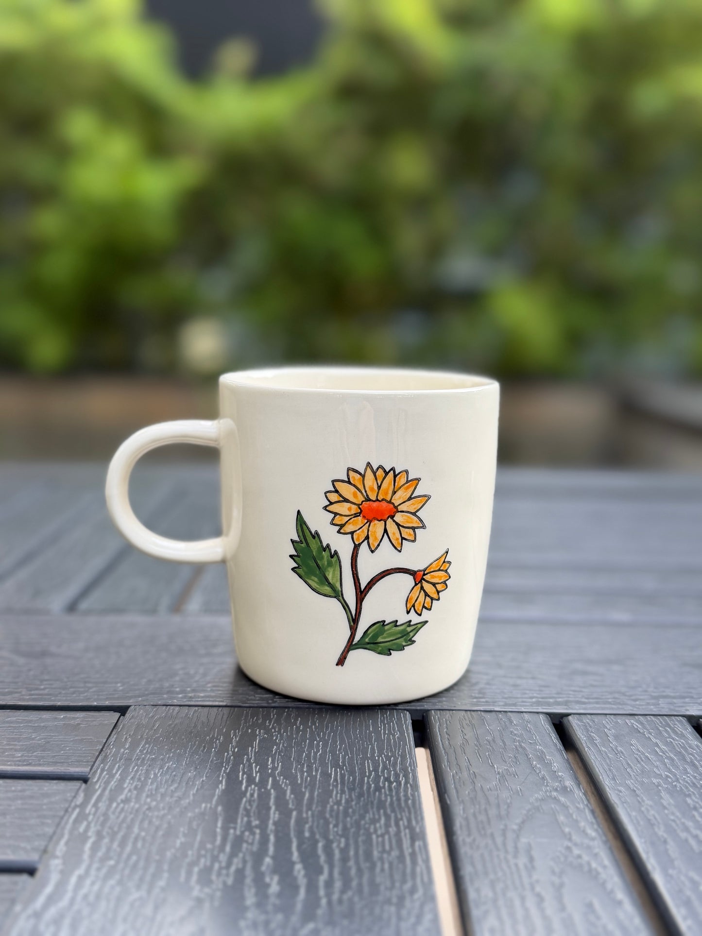 Hand-painted golden chrysanthemum ceramic mug, back view — one full bloom and one bud with orange centers and green leaves, handle on left, soft green backdrop
