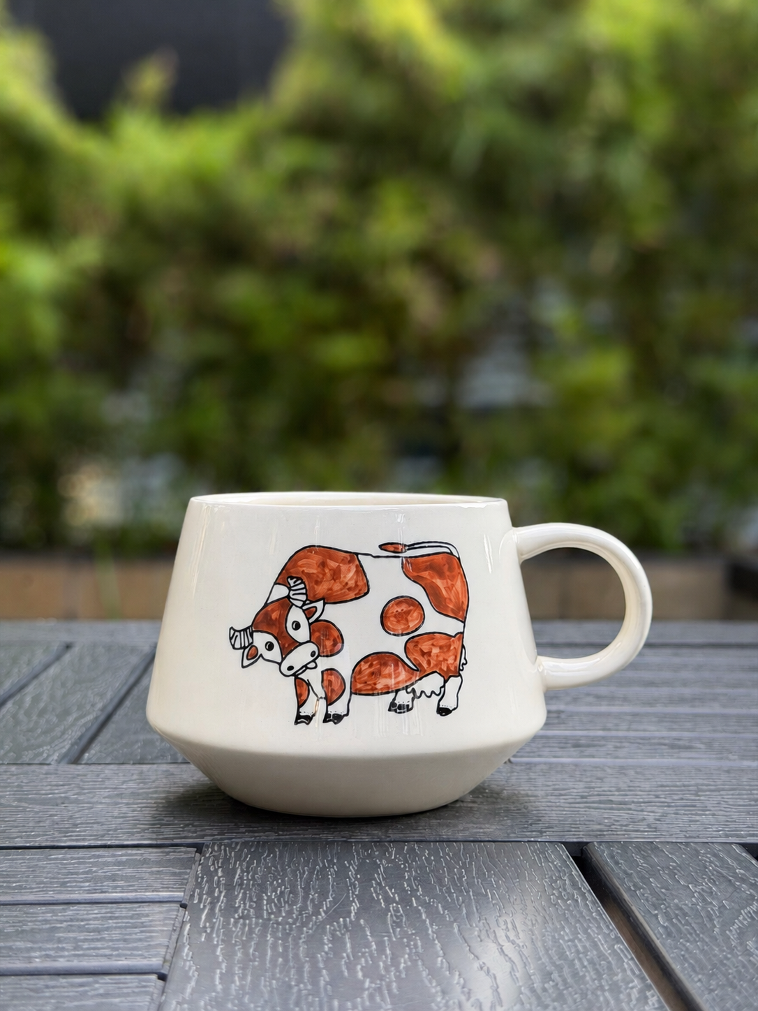 Hand-painted ceramic mug with brown cow illustration on cream glaze, placed on a wooden surface