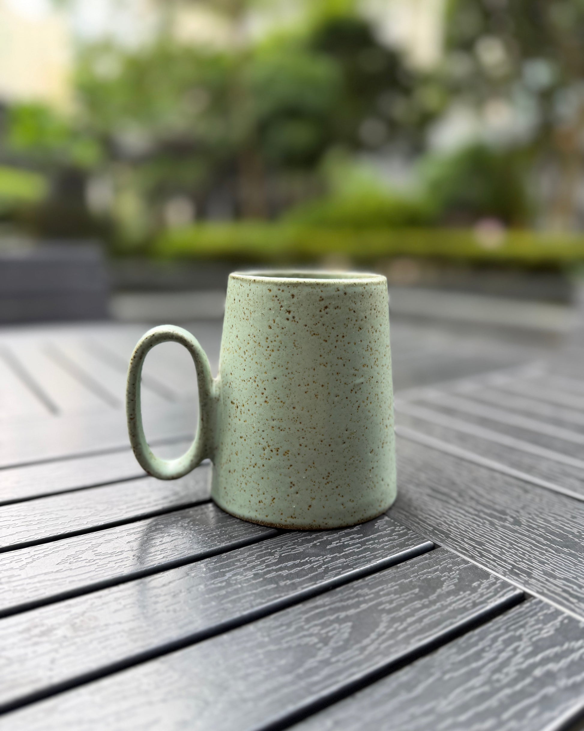 Quiet Meadow speckled sage green stoneware mug on a dark slatted outdoor table, handle facing left, showcasing the wide-base tapered silhouette and matte speckled glaze