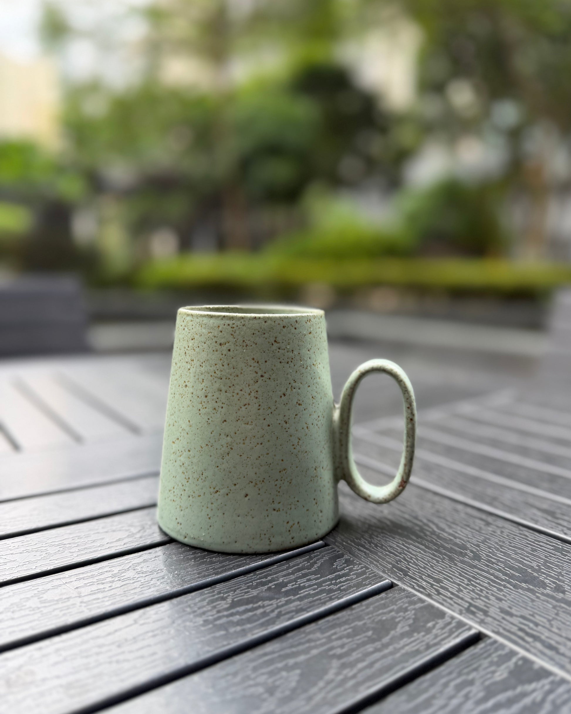 Quiet Meadow speckled sage green stoneware mug on a dark slatted outdoor table, handle facing right, with blurred green garden in the background