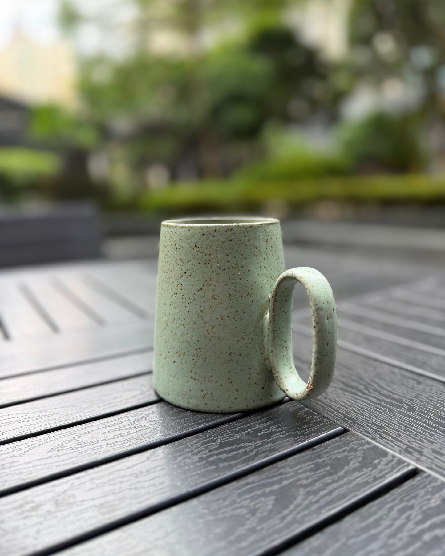 Side view of the Quiet Meadow speckled sage green stoneware mug on a dark slatted outdoor table, highlighting the oval loop handle and tapered body with natural speckle texture
