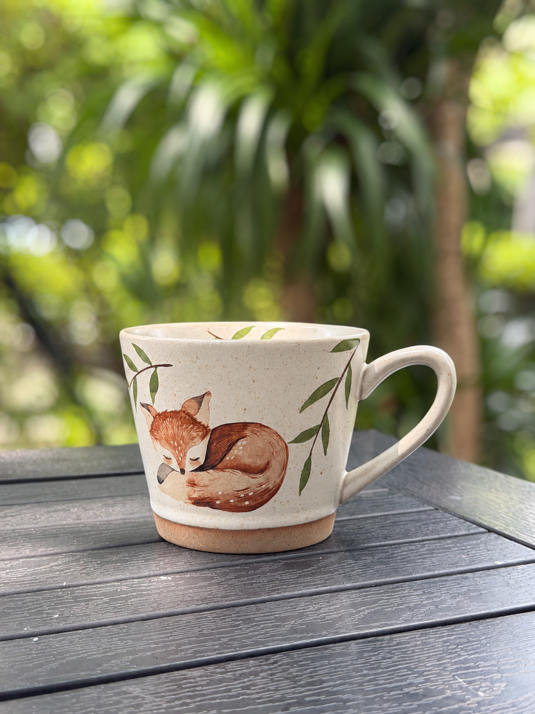 Sleeping fox ceramic mug on a dark outdoor table with greenery in the background