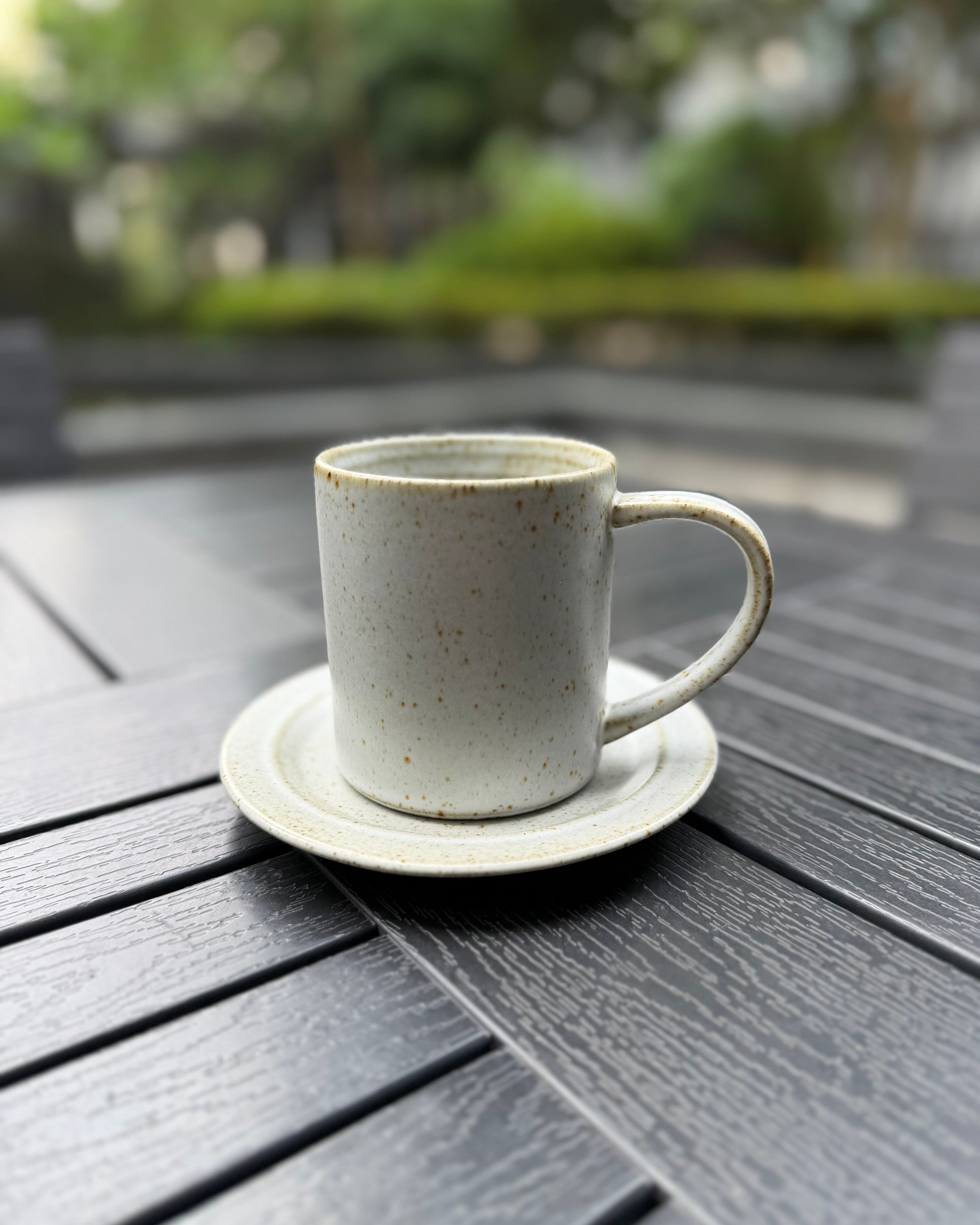 A speckled off-white ceramic coffee mug with a matching saucer, placed on a dark gray outdoor wooden table with a blurred garden background.