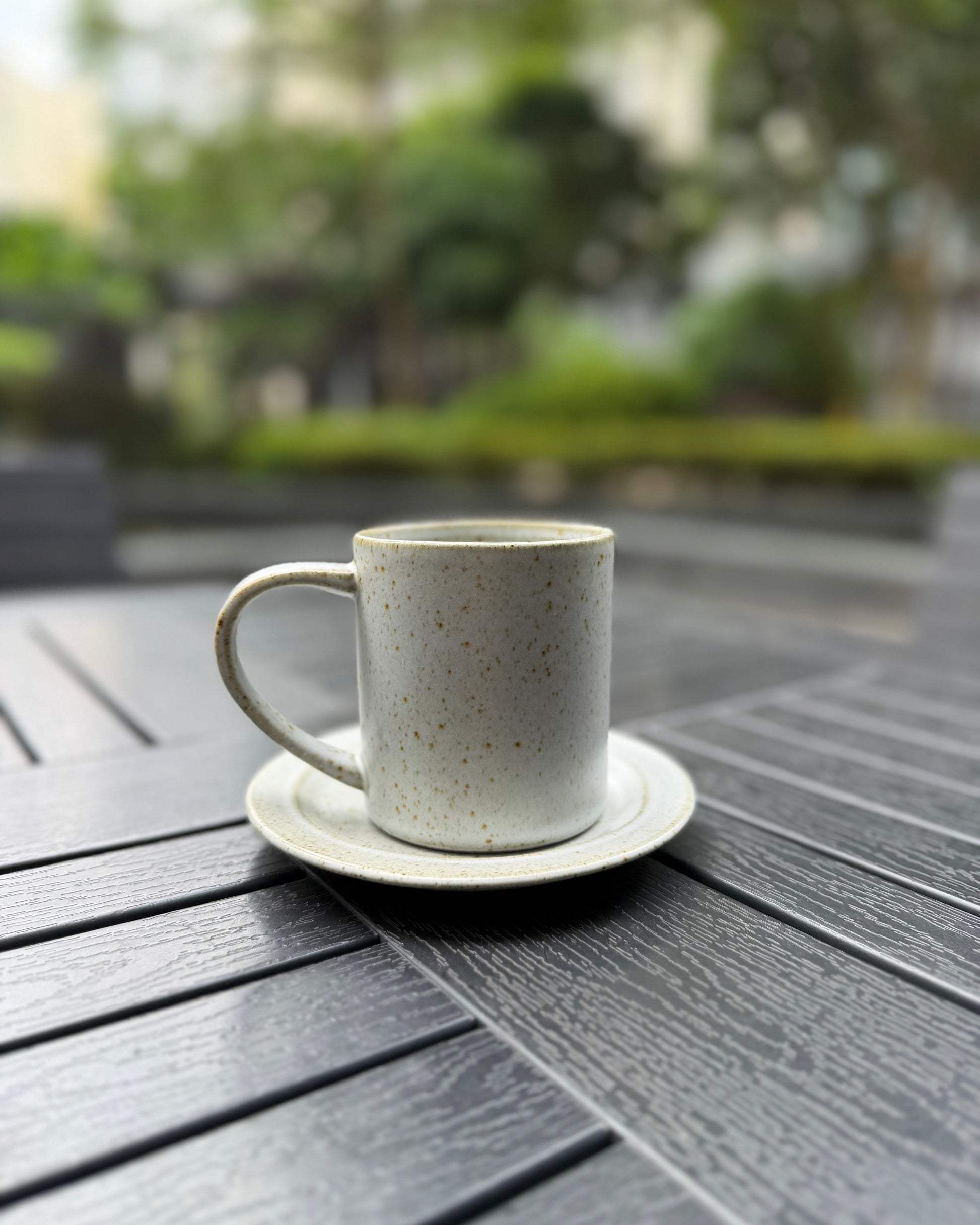 A speckled ceramic coffee mug with a matching saucer, placed on a dark gray slatted outdoor table with a blurred garden background.