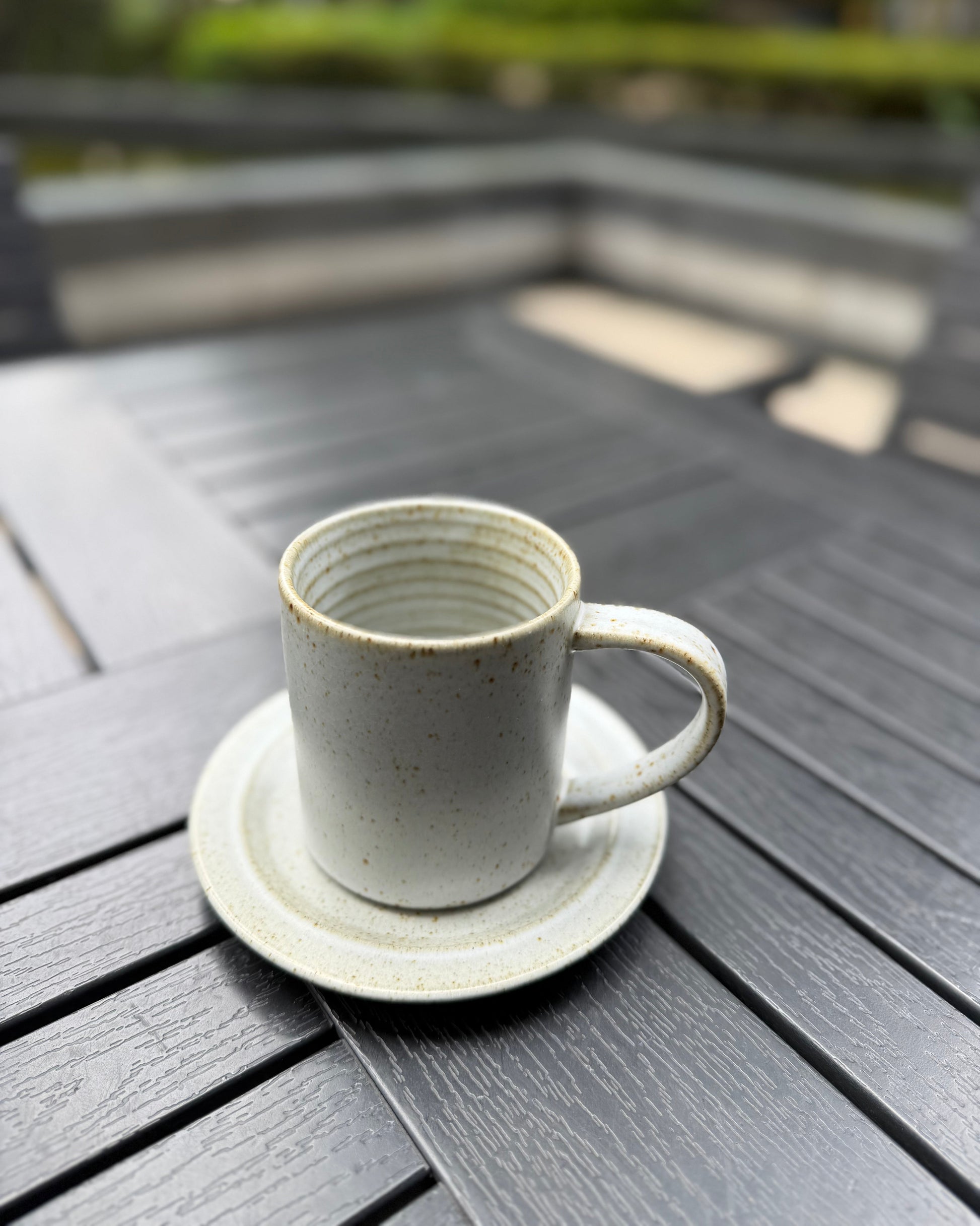 A small speckled ceramic cup with a matching saucer sits on a dark gray outdoor table, with a blurred garden background.